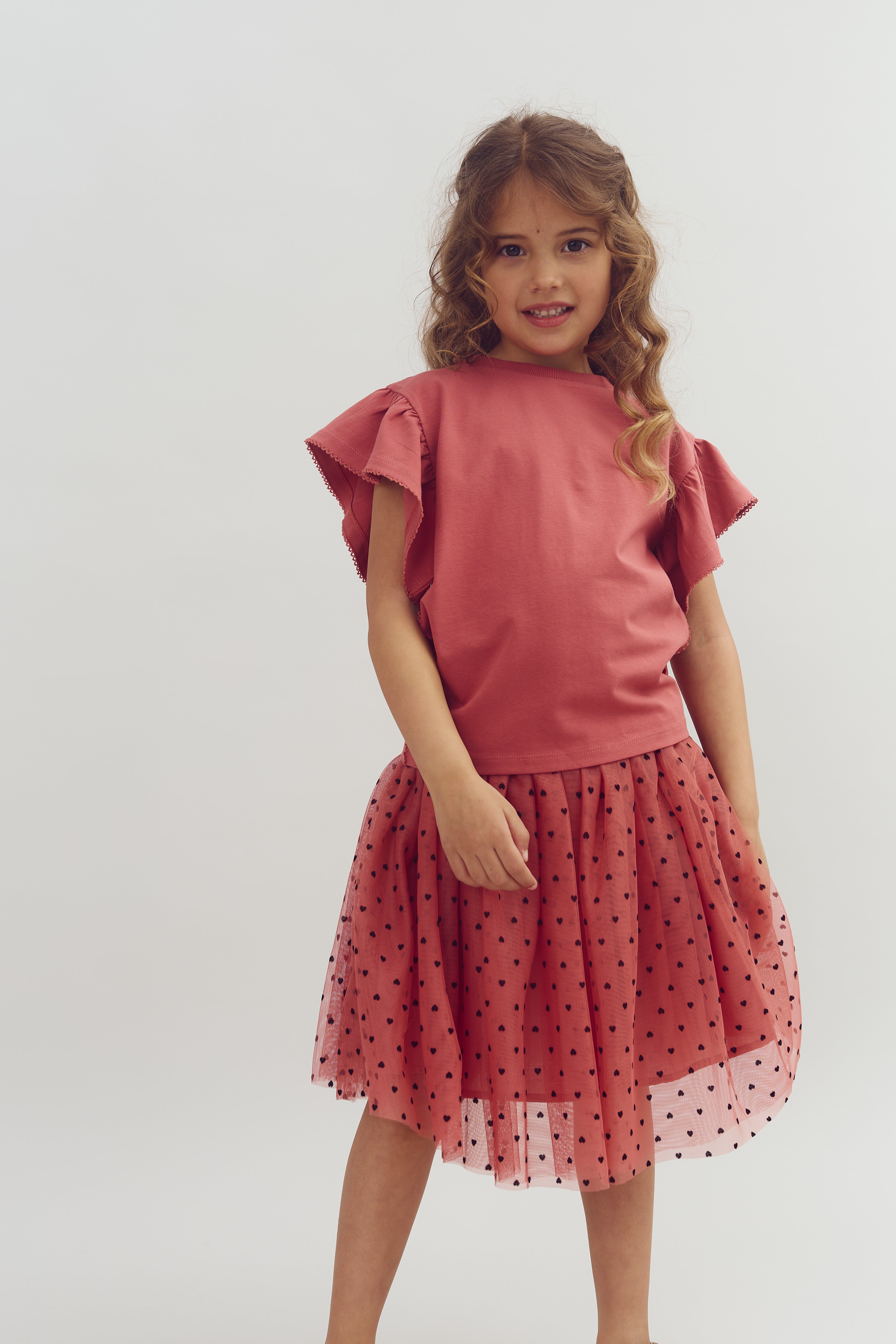 A child stands smiling, wearing a pink top with ruffled sleeves and a matching polka-dotted tulle skirt. The background is a simple, plain white studio setting.