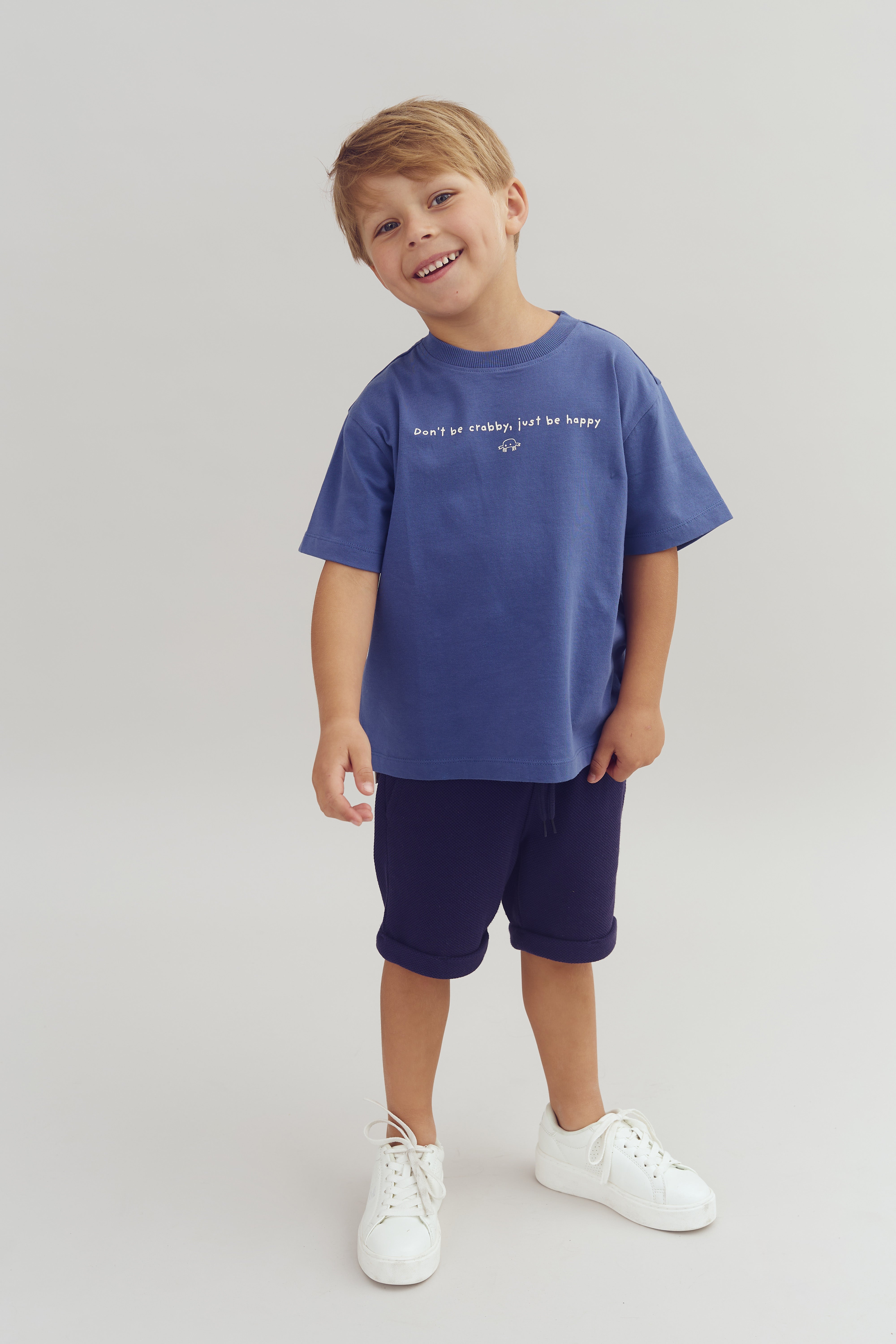 A smiling child stands wearing a blue T-shirt with the text "Don't be crabby, just be happy," navy shorts, and white sneakers against a plain background.