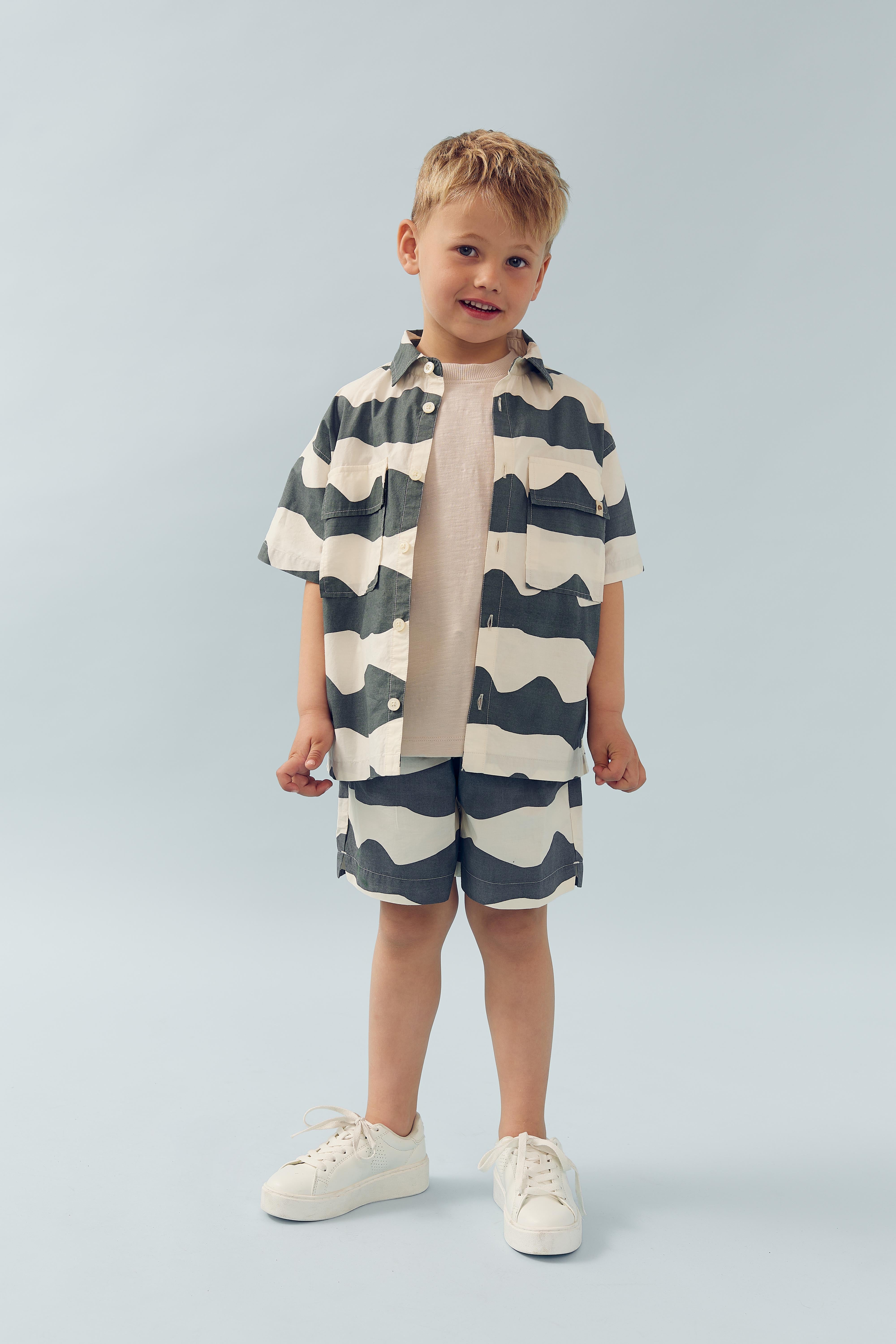 A young child stands smiling, wearing a short-sleeved, striped shirt and matching shorts, with white sneakers. The background is a plain light blue.