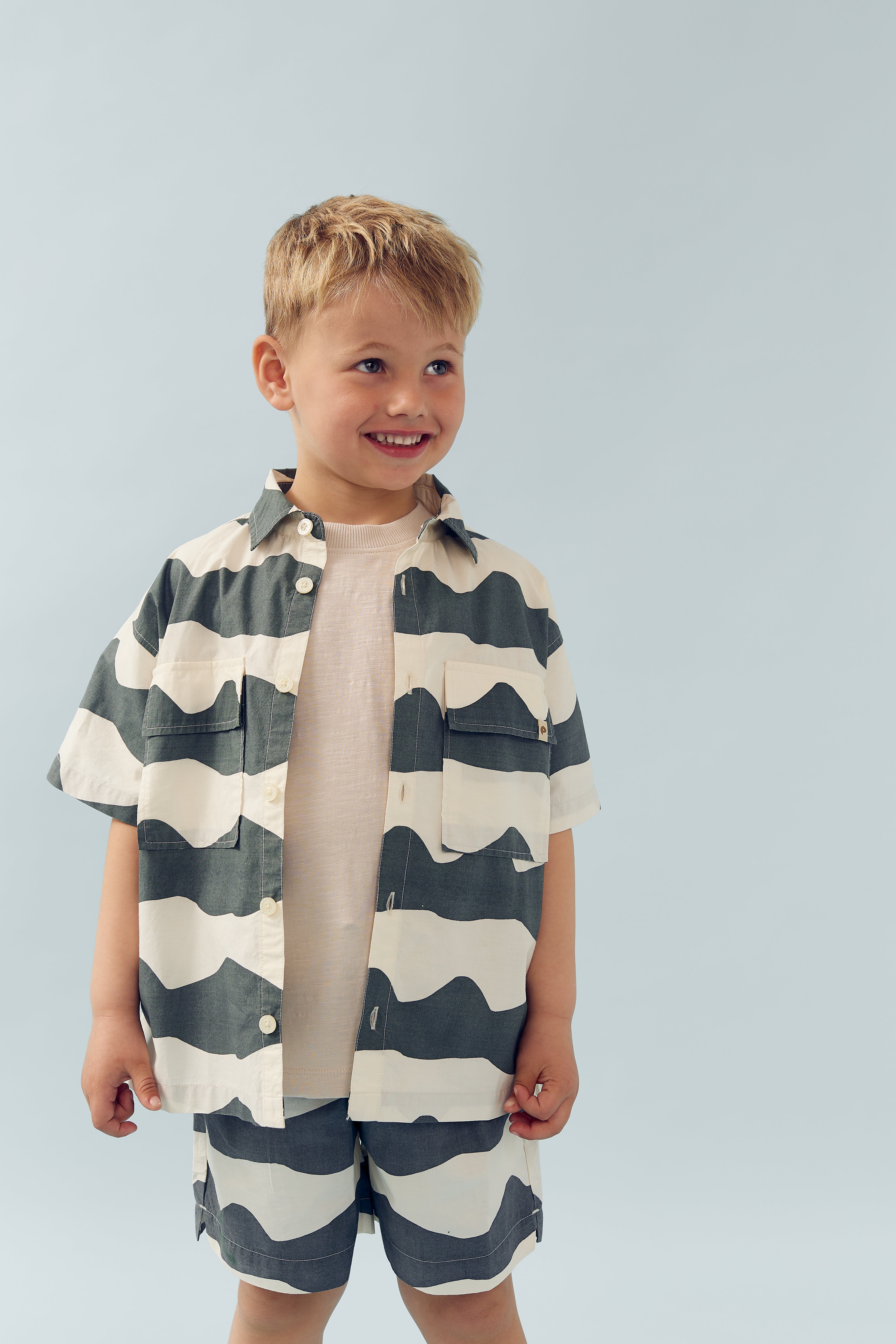 A young child, smiling, wears a black-and-white wavy striped shirt and matching shorts, standing against a plain light blue background.