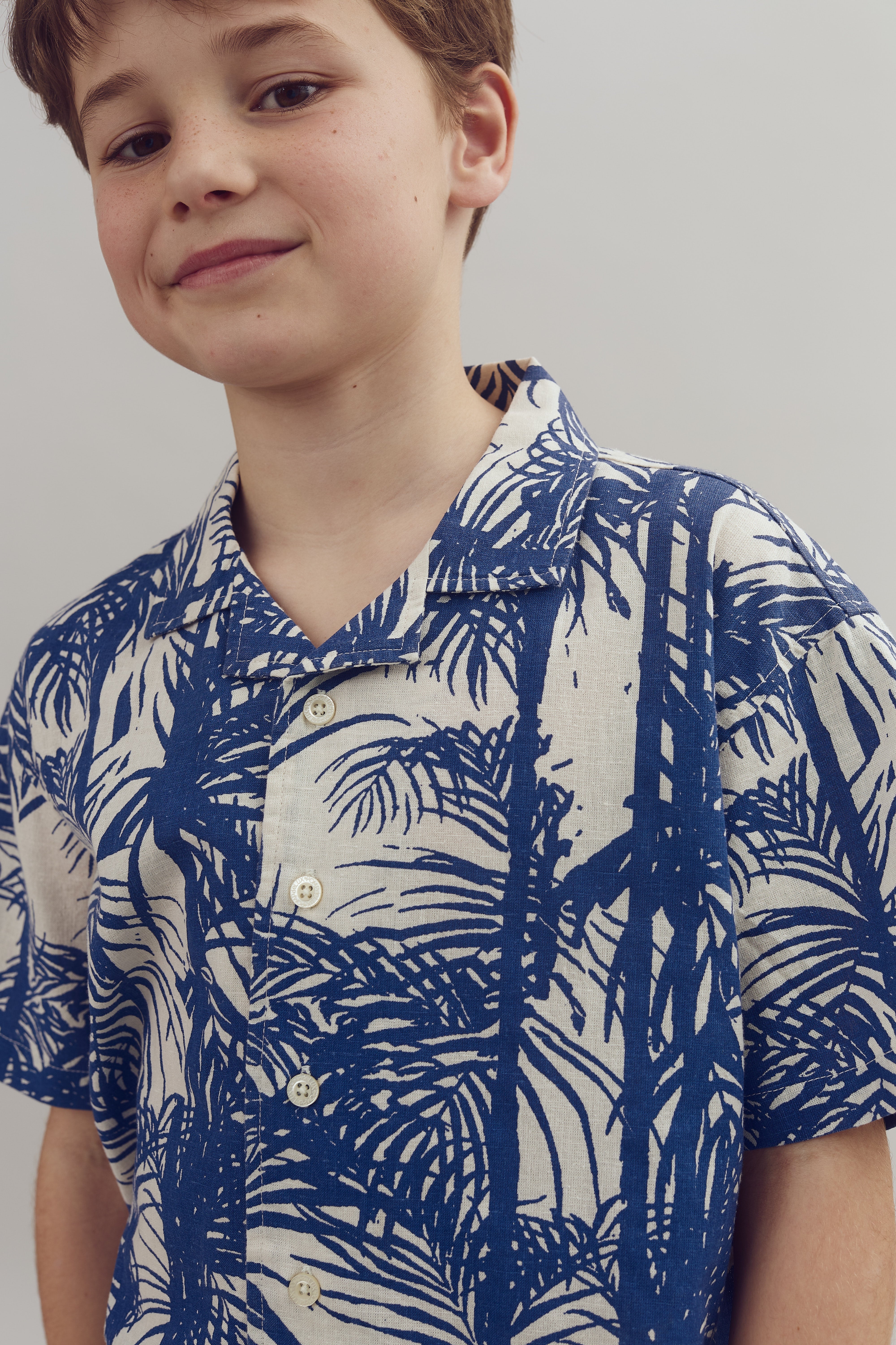 A young boy smiles subtly, wearing a button-up shirt with a blue and white palm tree pattern. The background is a plain, light-colored wall.