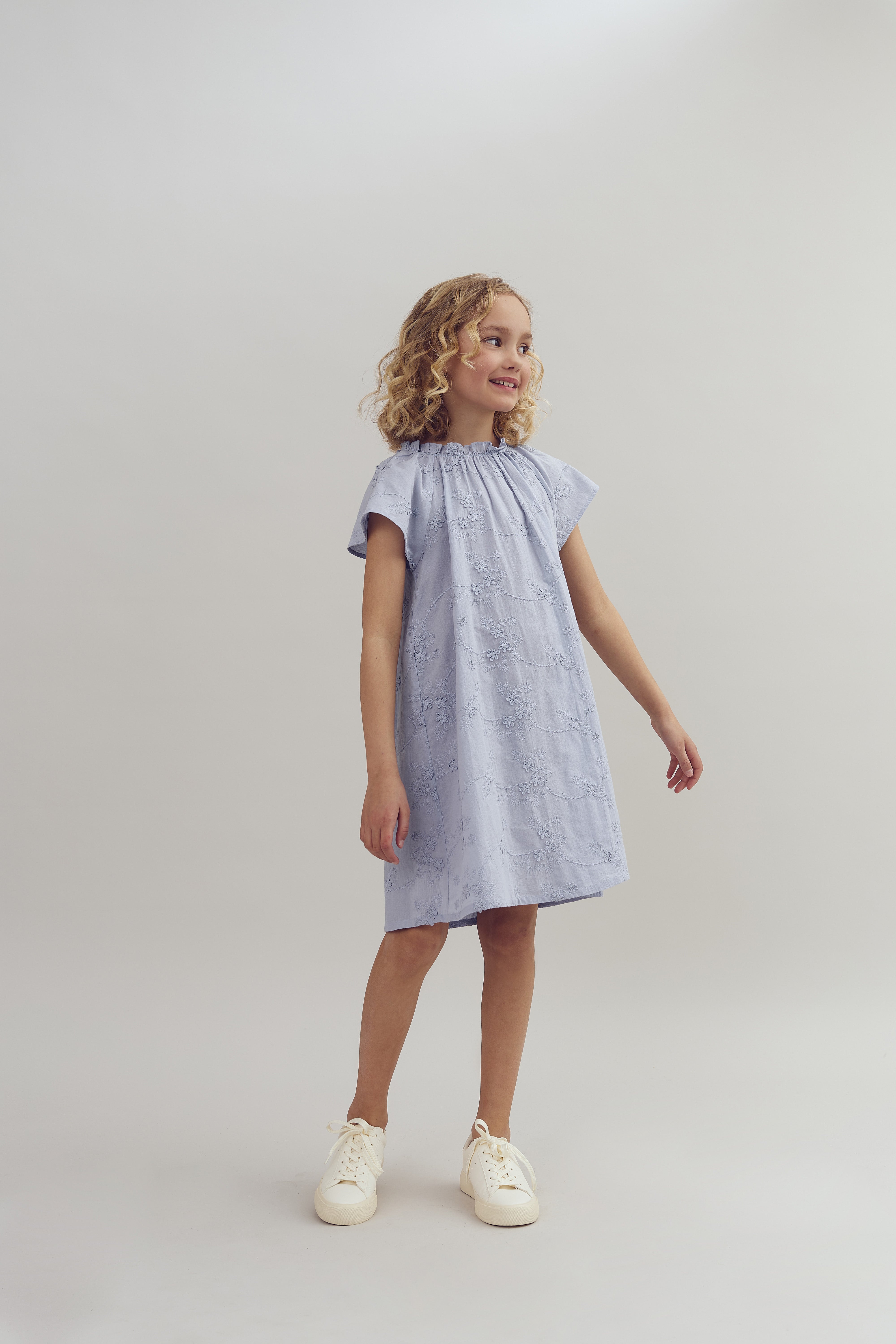 A young girl in a light blue dress stands smiling with arms slightly extended in a neutral, light-colored studio. She wears white sneakers, and her hair is curly.