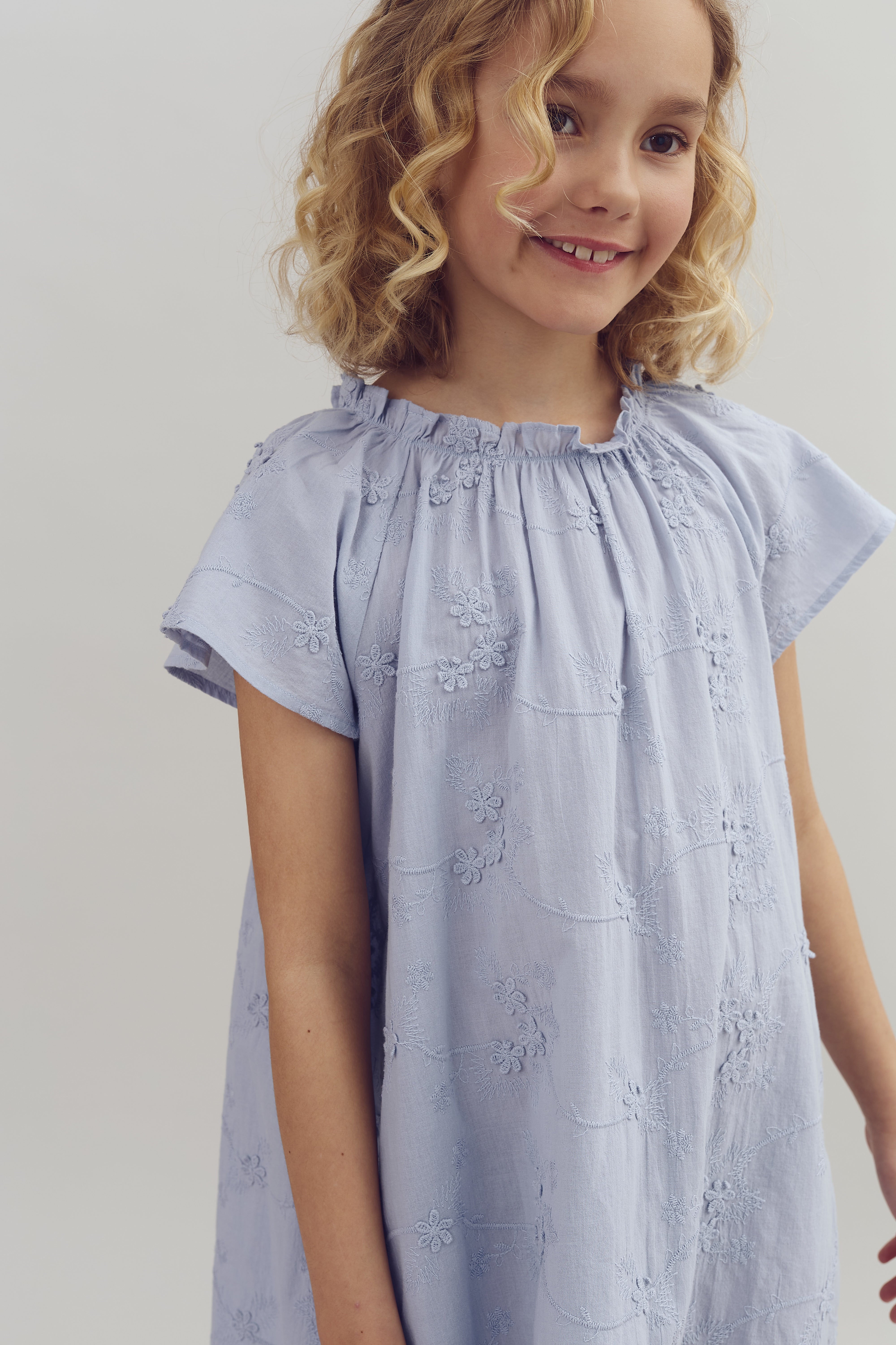 A young girl with curly hair smiles gently, wearing a light blue, floral-embroidered dress, standing against a plain, neutral background.