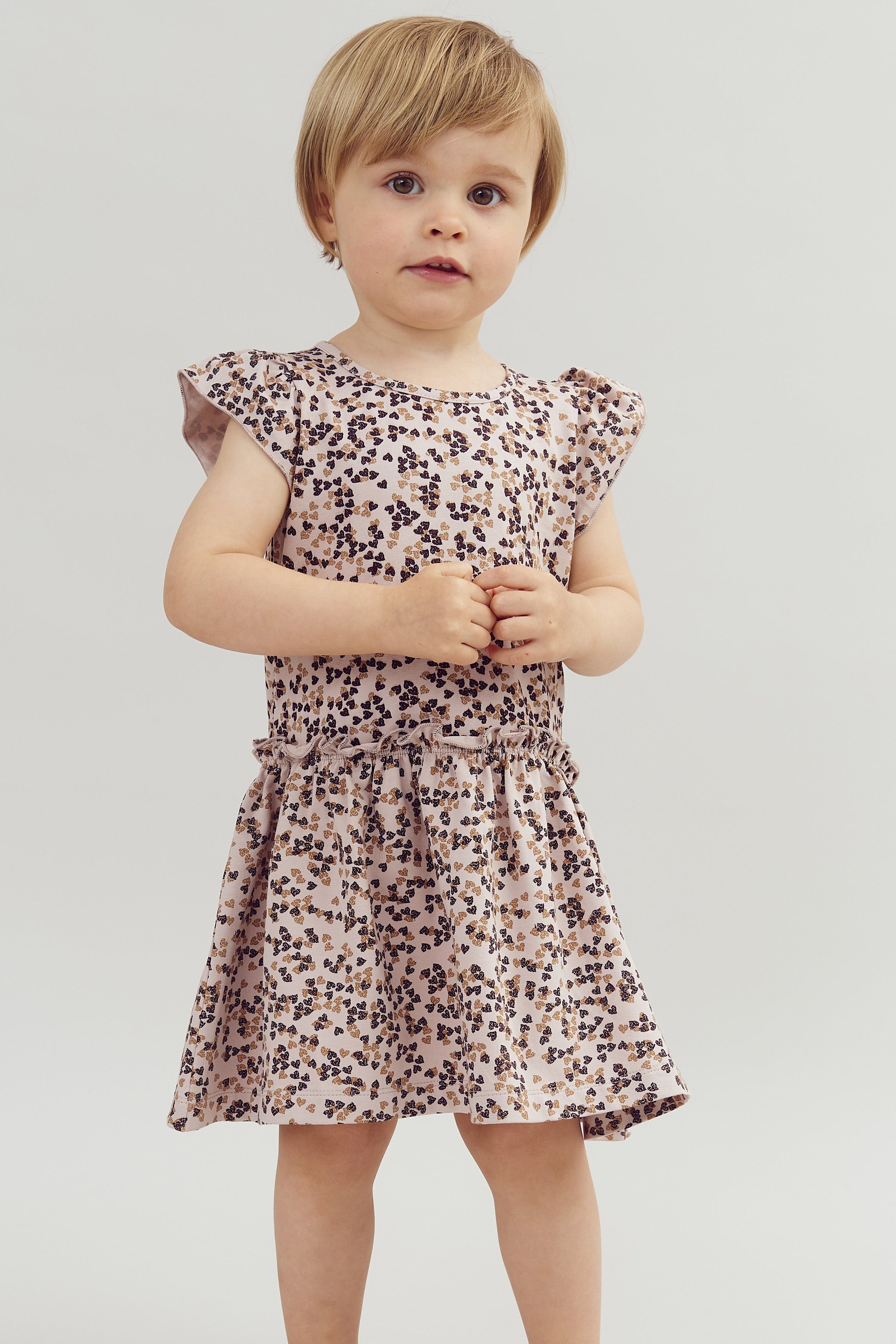 A young child stands wearing a patterned dress and brown shoes, hands clasped. The background is plain, white, and minimalistic.