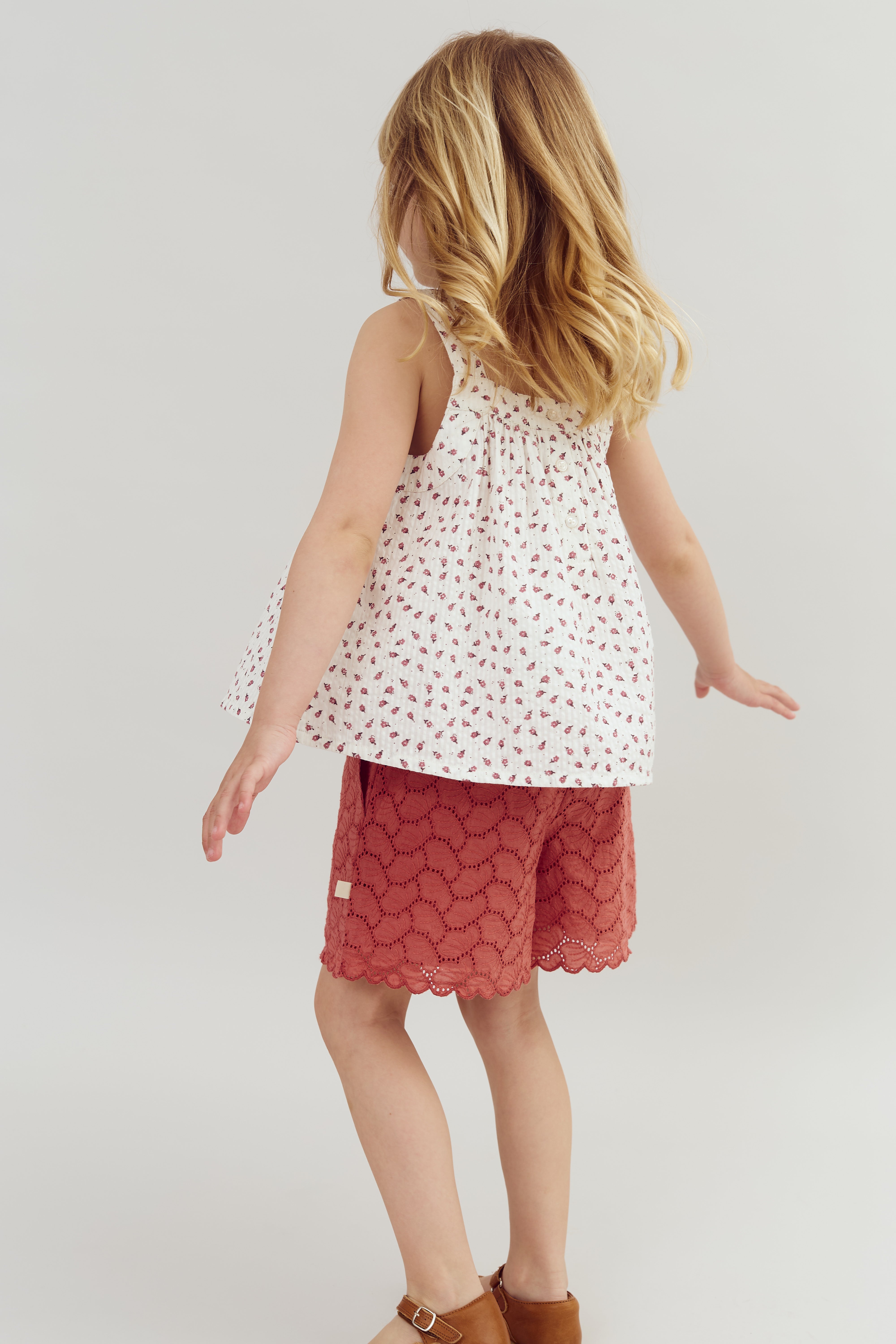 A child facing away, wearing a floral top and red eyelet shorts, stands with arms slightly outstretched in a neutral, light-colored setting.