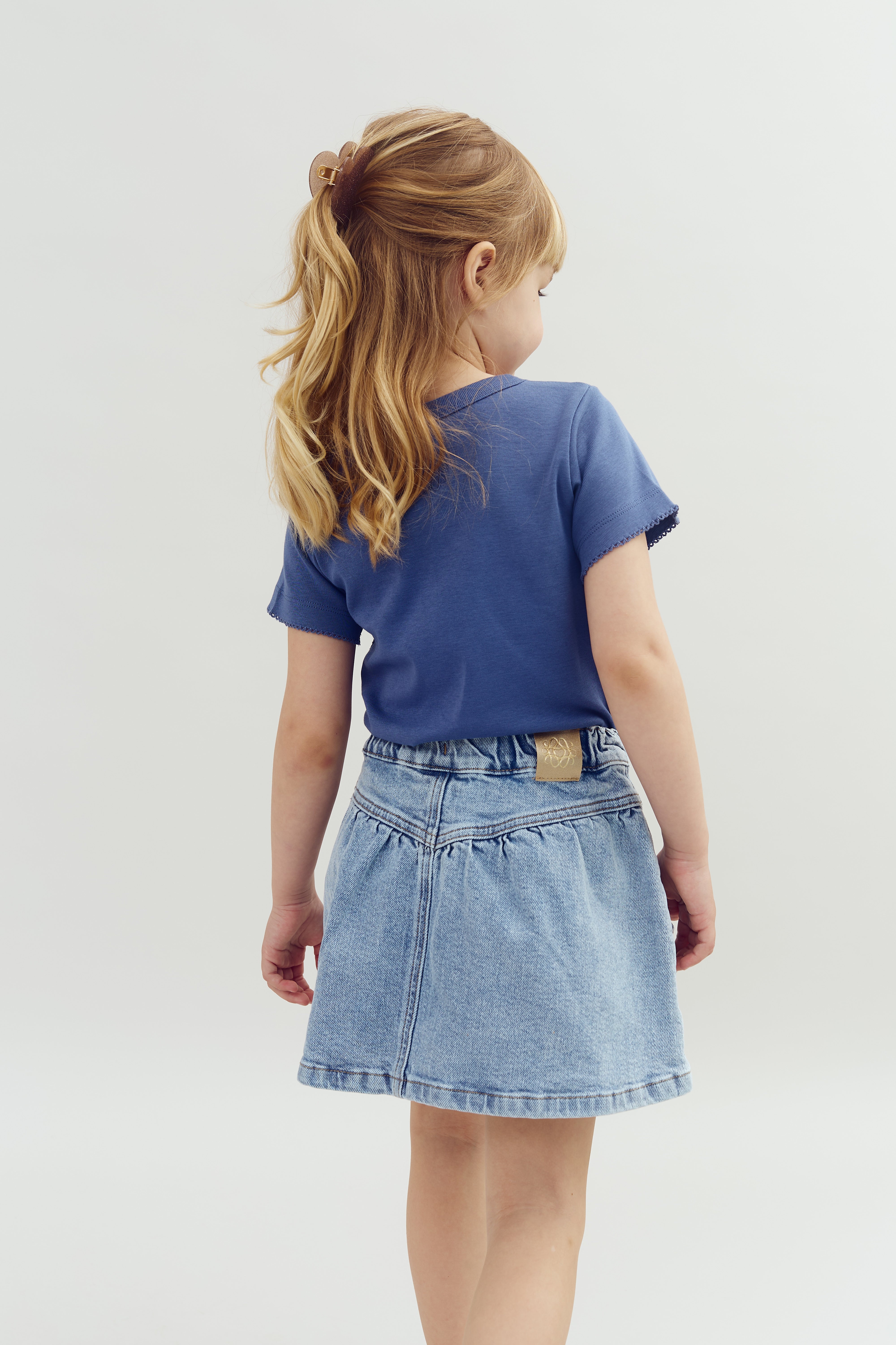 A young girl with blonde hair wears a blue shirt and denim skirt, facing away in a neutral, light-colored setting.