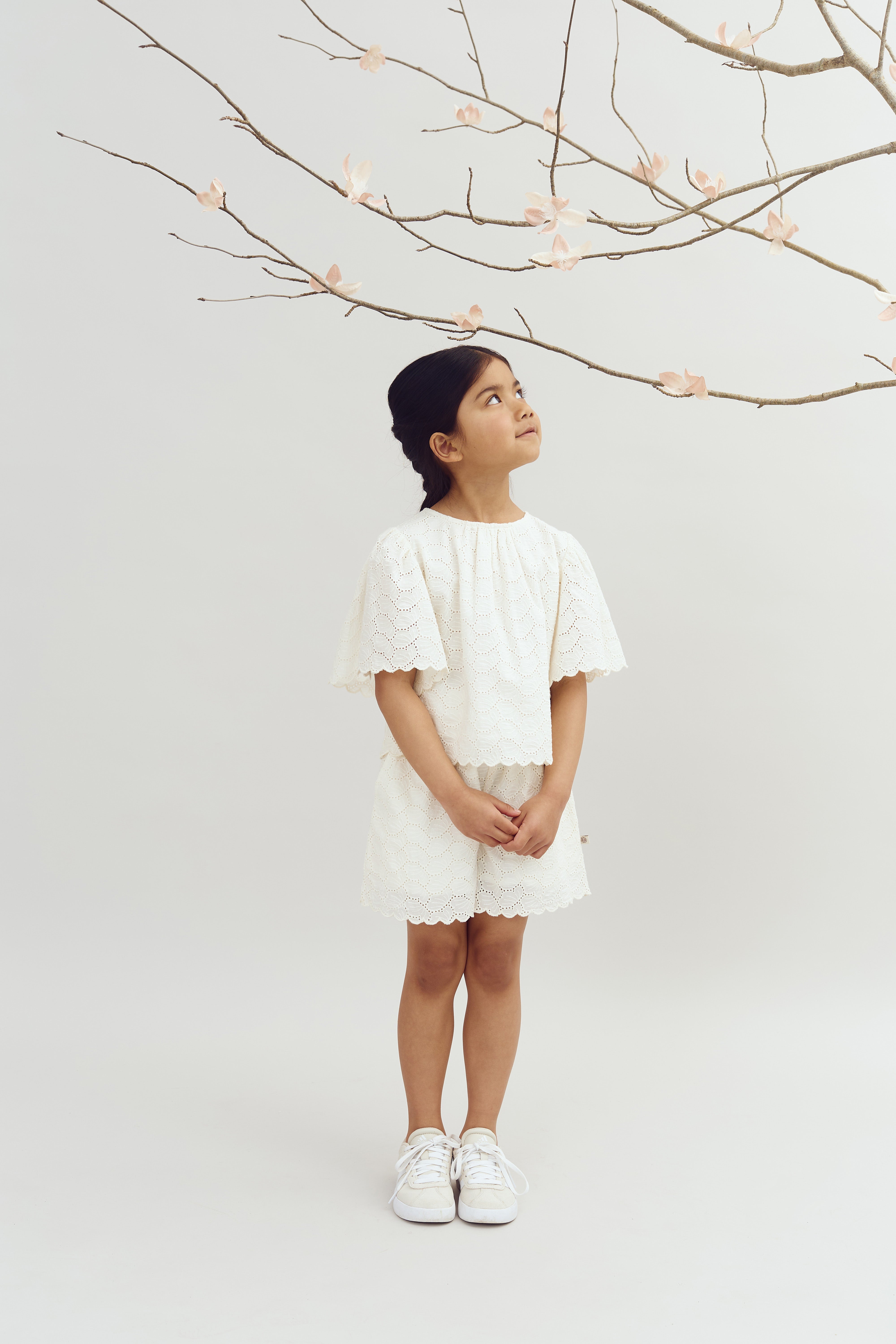 A young girl stands wearing a white dress and sneakers, gazing up at a bare branch adorned with pink blossoms, against a plain light background.