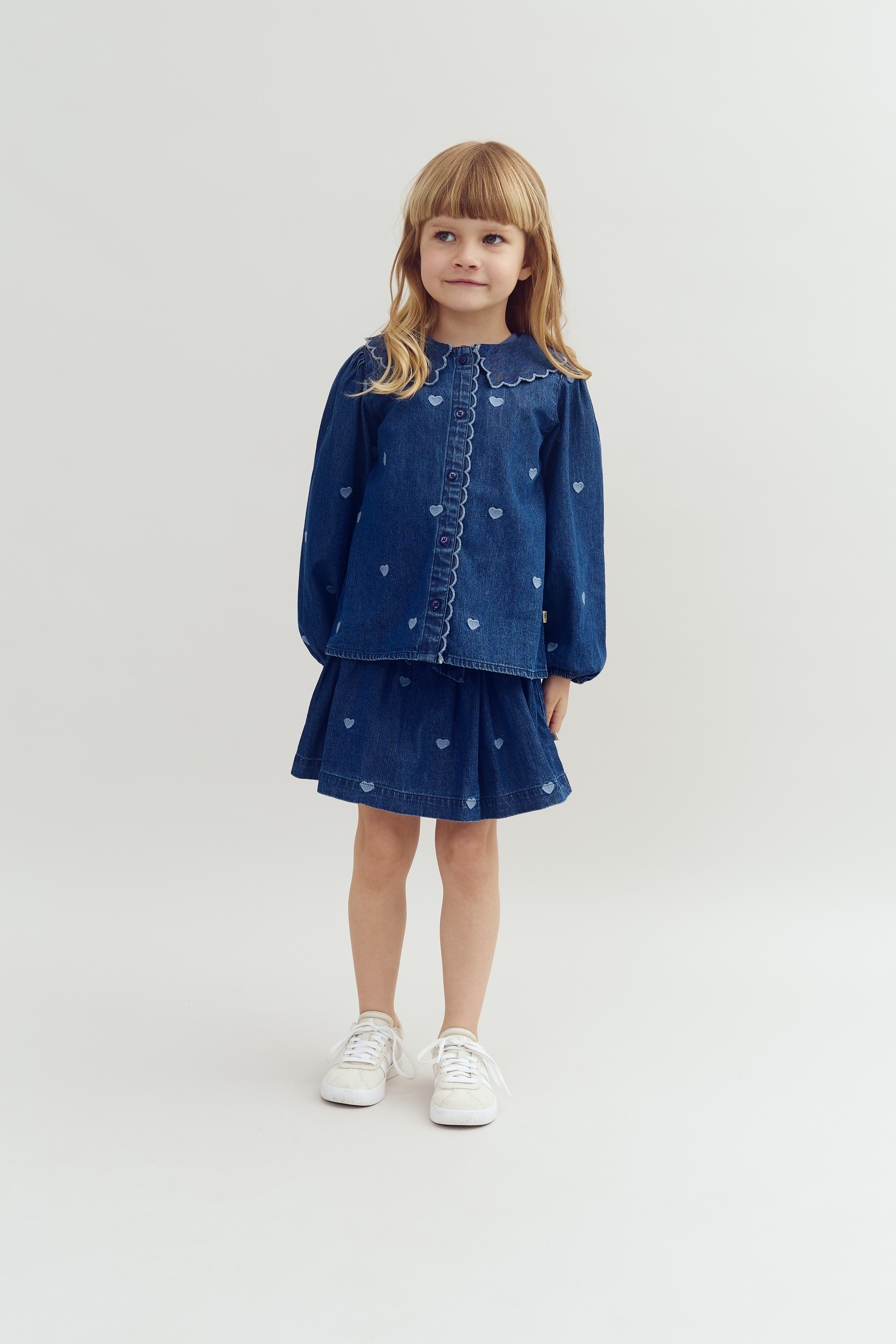 A young girl stands, wearing a matching denim shirt and skirt with heart patterns and white sneakers, against a plain light-colored background.