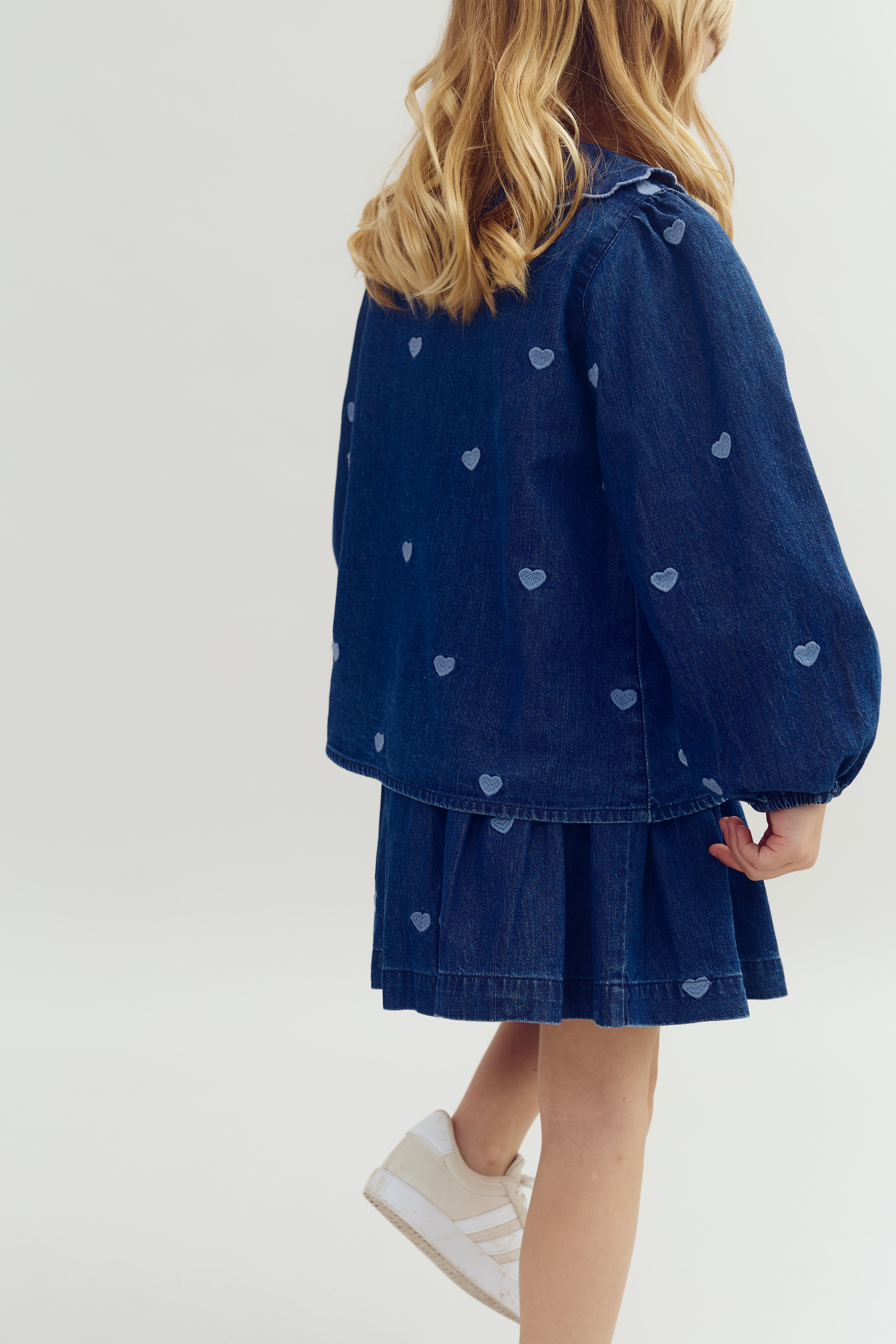 A child wearing a blue denim jacket and skirt adorned with small heart patterns is standing, facing away. Their blond hair falls over the jacket, and they wear white sneakers.