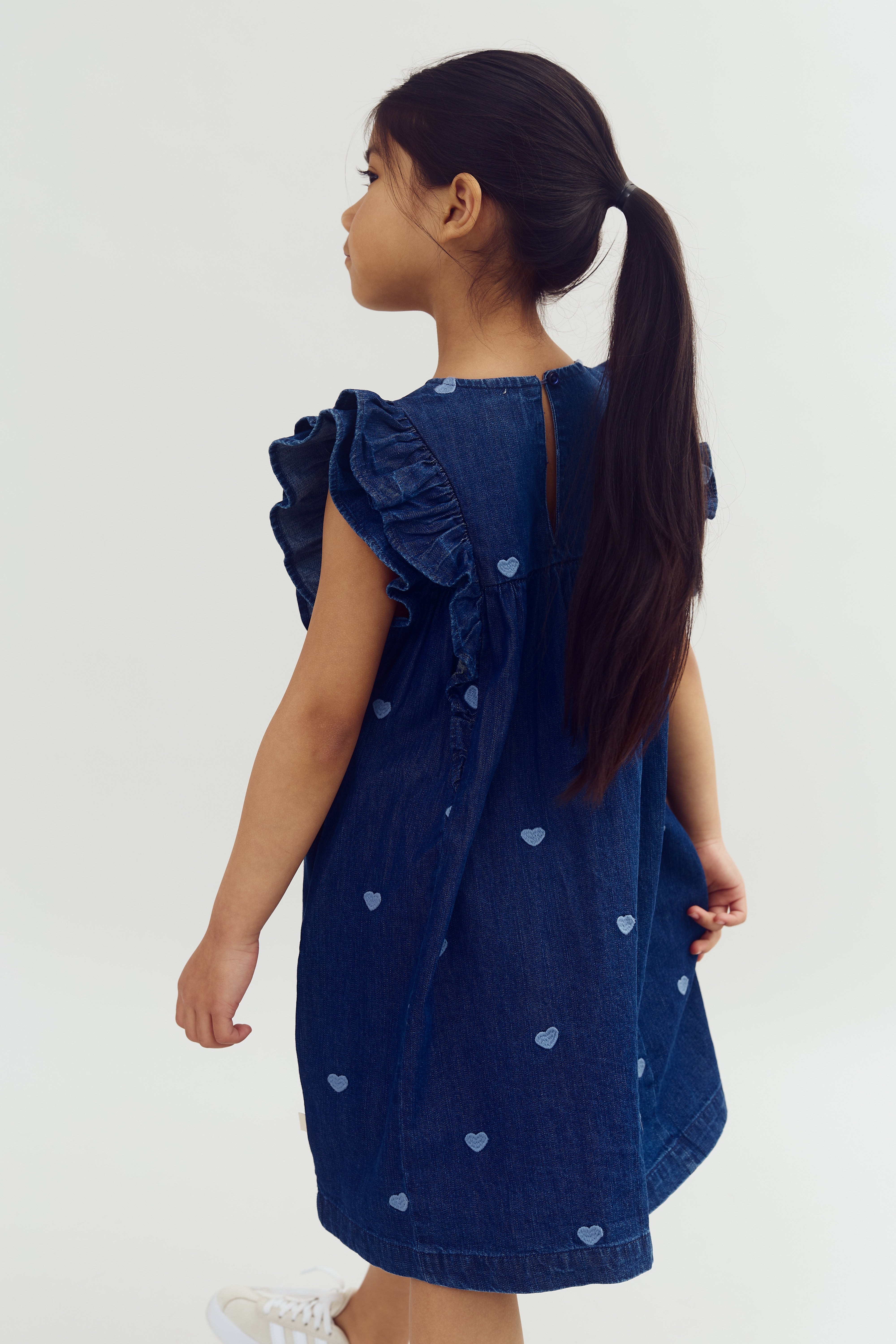 A child with long hair tied back wears a denim dress with ruffled sleeves and heart patterns, standing against a plain background.