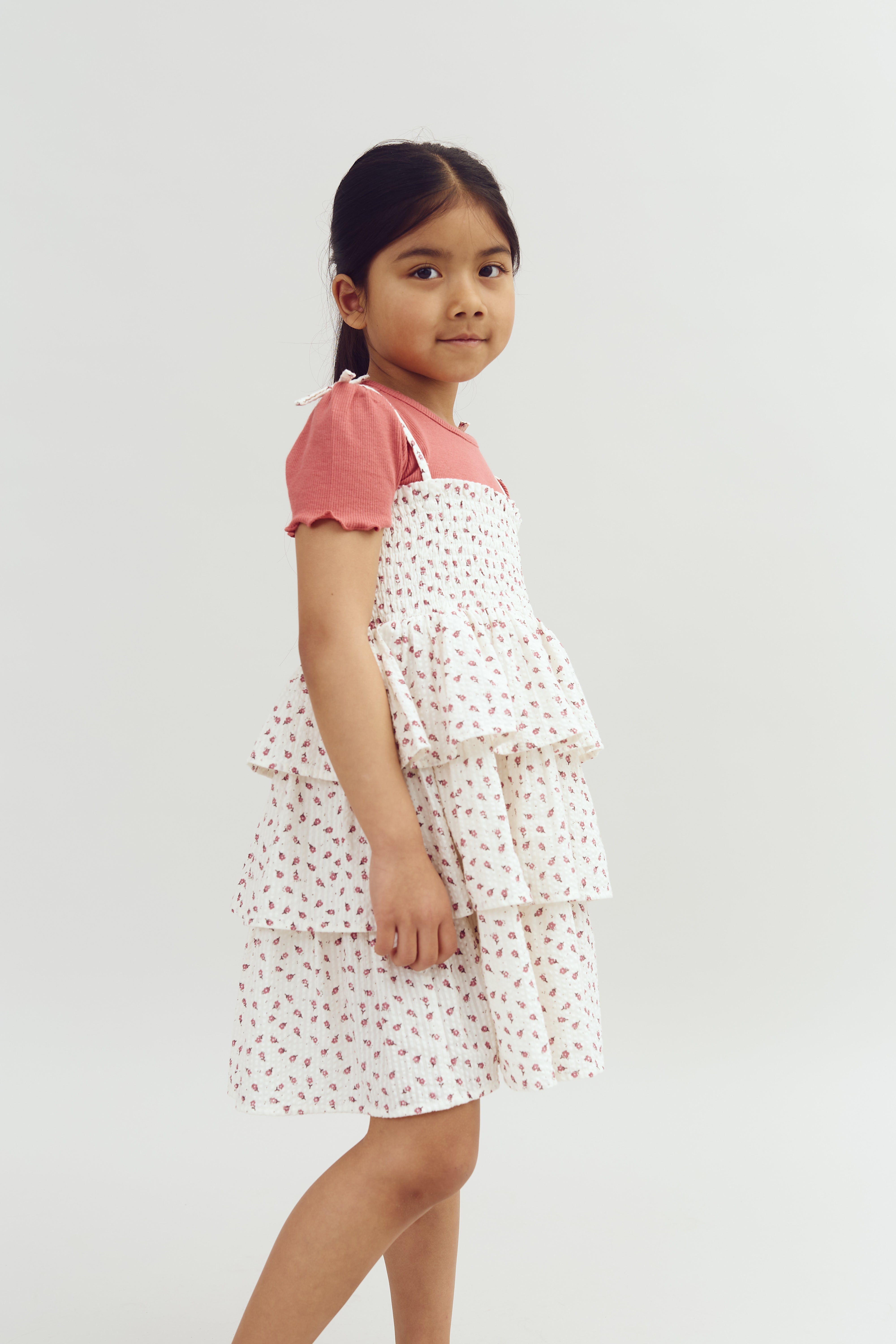 A young girl looks at the camera, wearing a pink top with a white, floral tiered dress, posing against a plain white background.