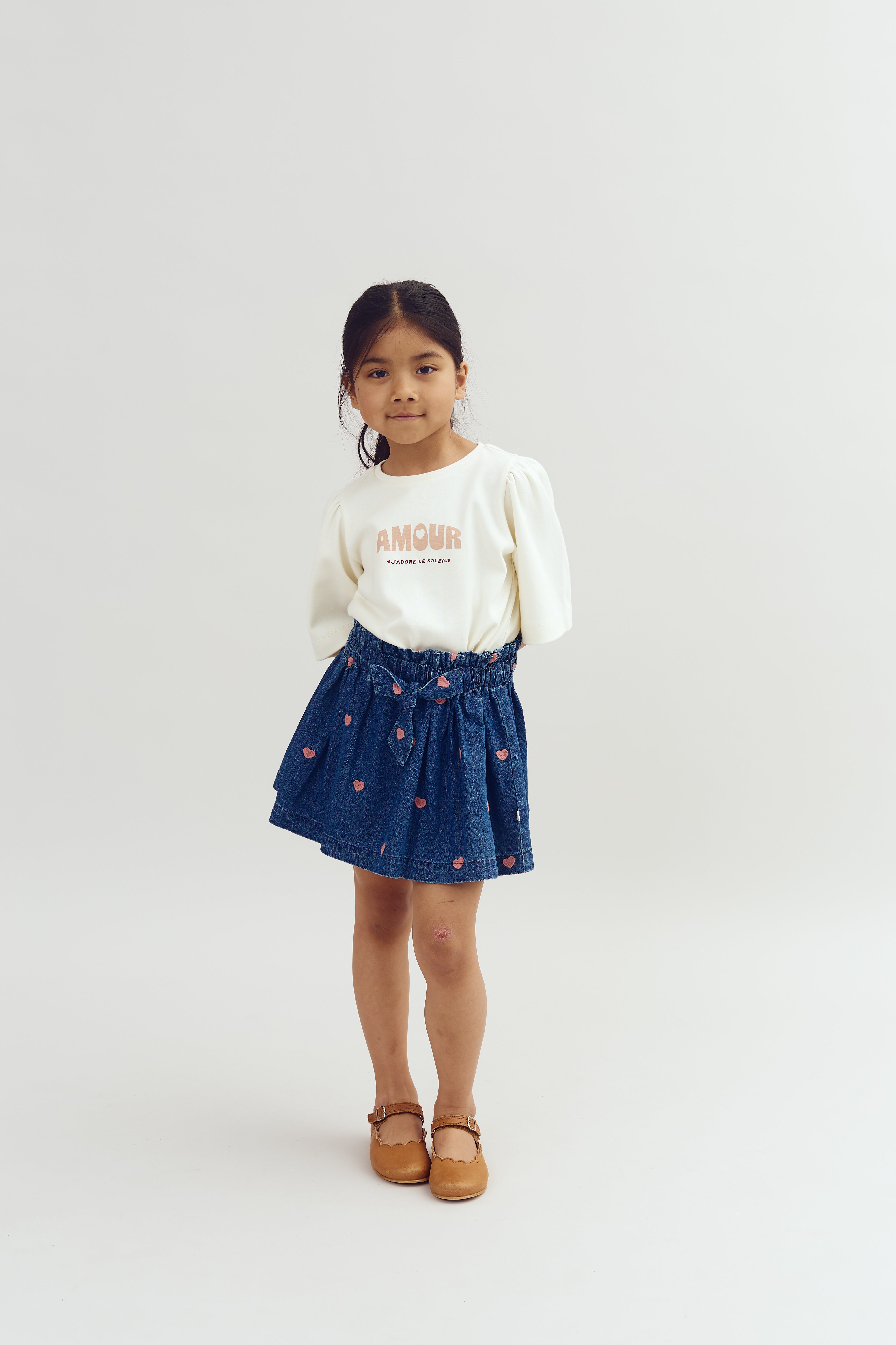 A young girl stands facing forward. She wears a white sweater with "AMOUR" and "J’adore le soleil," a blue skirt with pink hearts, and brown shoes, against a plain background.