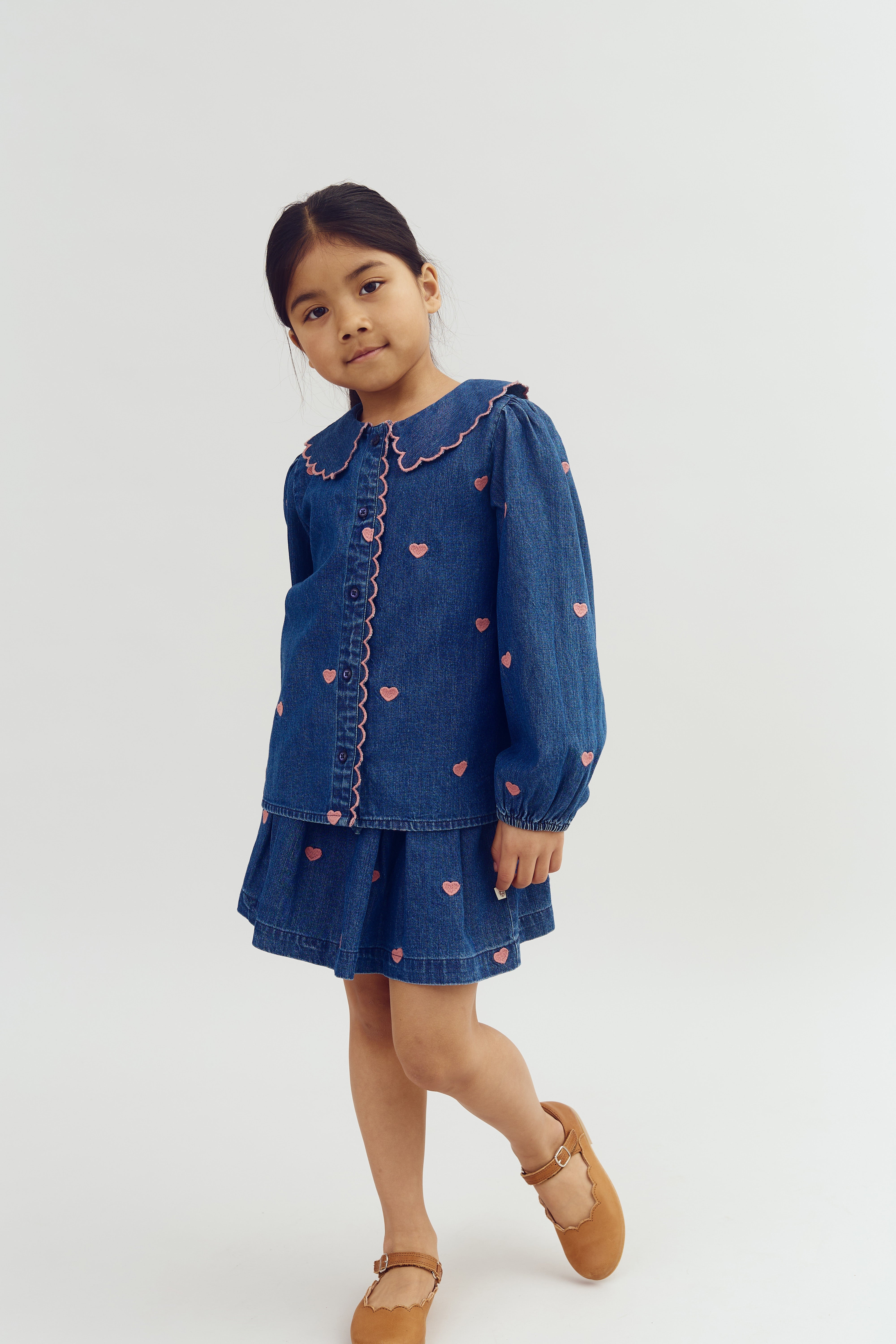 A child wearing a denim dress adorned with pink hearts and scalloped edges stands against a plain, light background, with one leg slightly bent, creating a playful appearance.