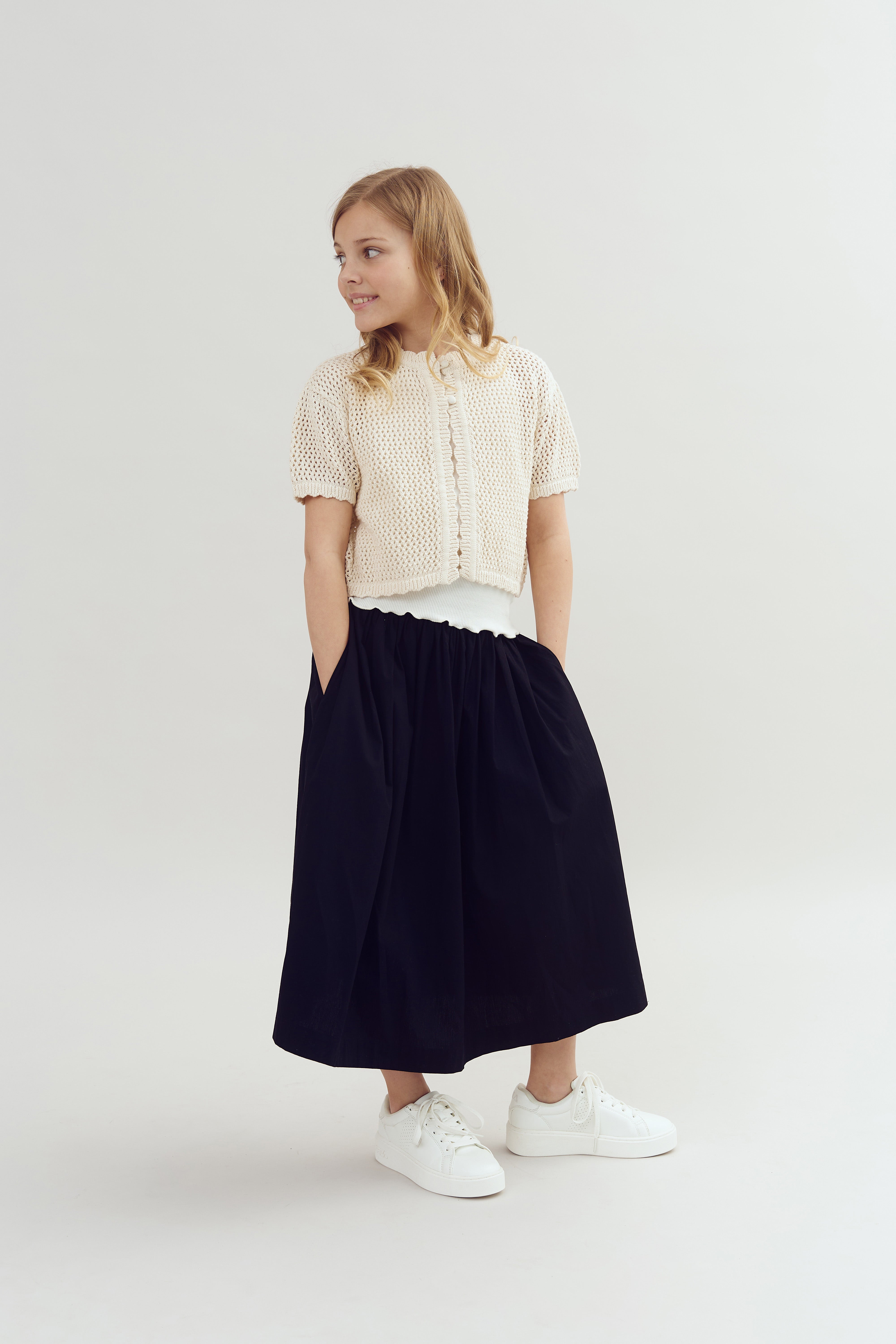A young girl stands smiling, hands in pockets, wearing a cream knitted cardigan, black skirt, and white sneakers, against a plain light-colored background.
