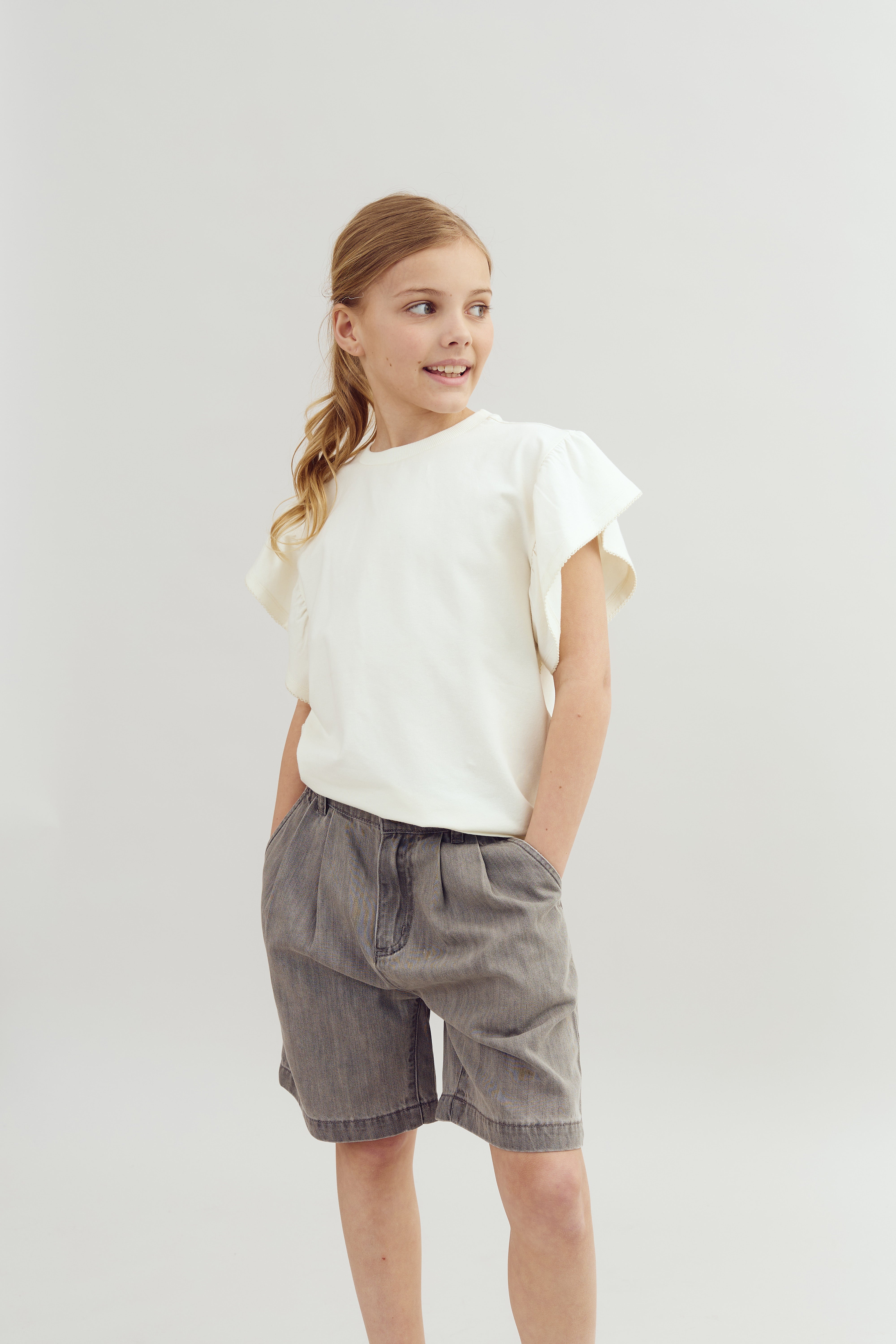 A young girl stands with hands in pockets, wearing a white shirt and gray shorts, looking sideways in a bright, neutral space.