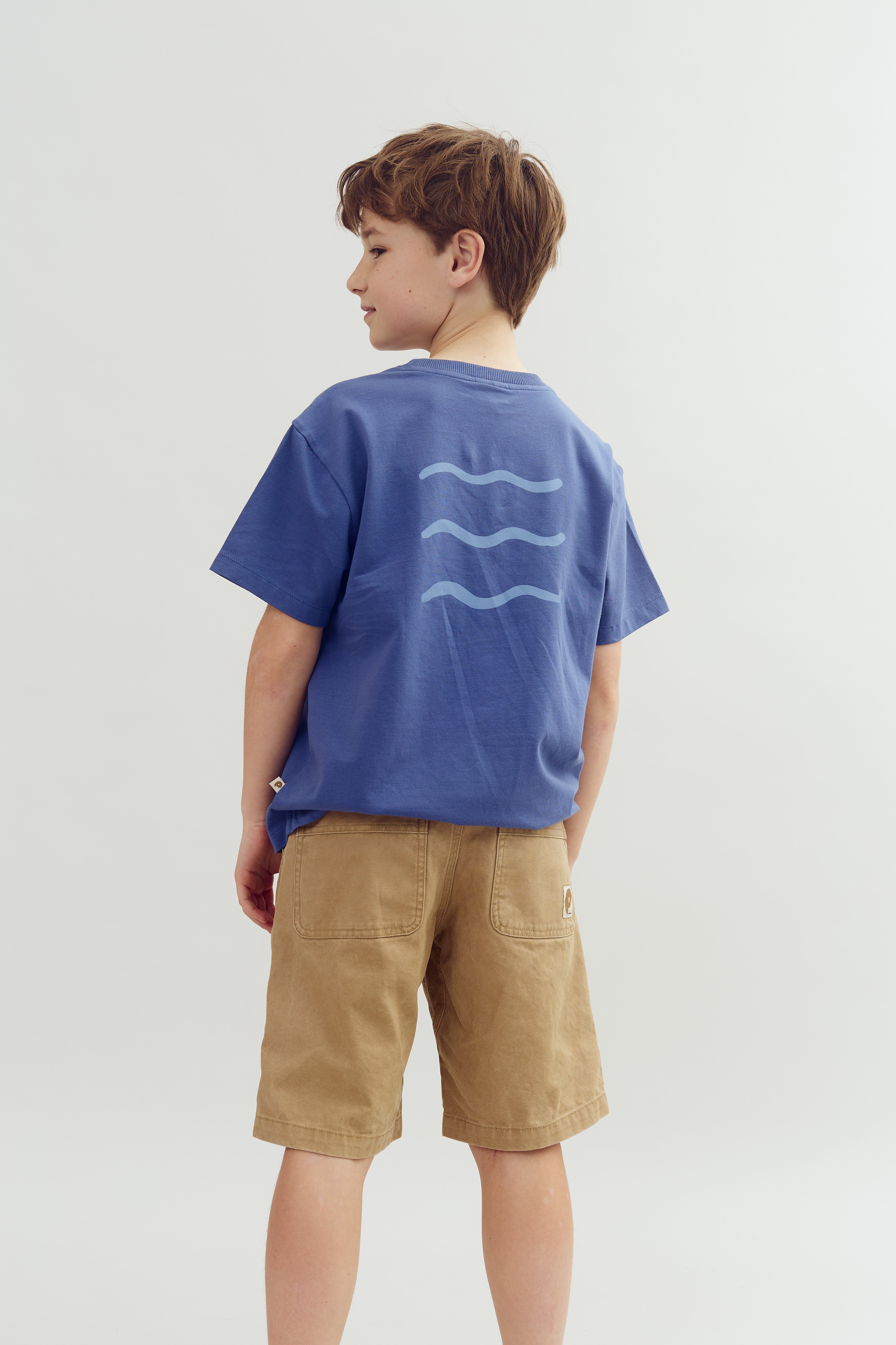 A boy stands turned to the side, wearing a blue T-shirt with three wave symbols on the back and brown shorts. The background is plain white.