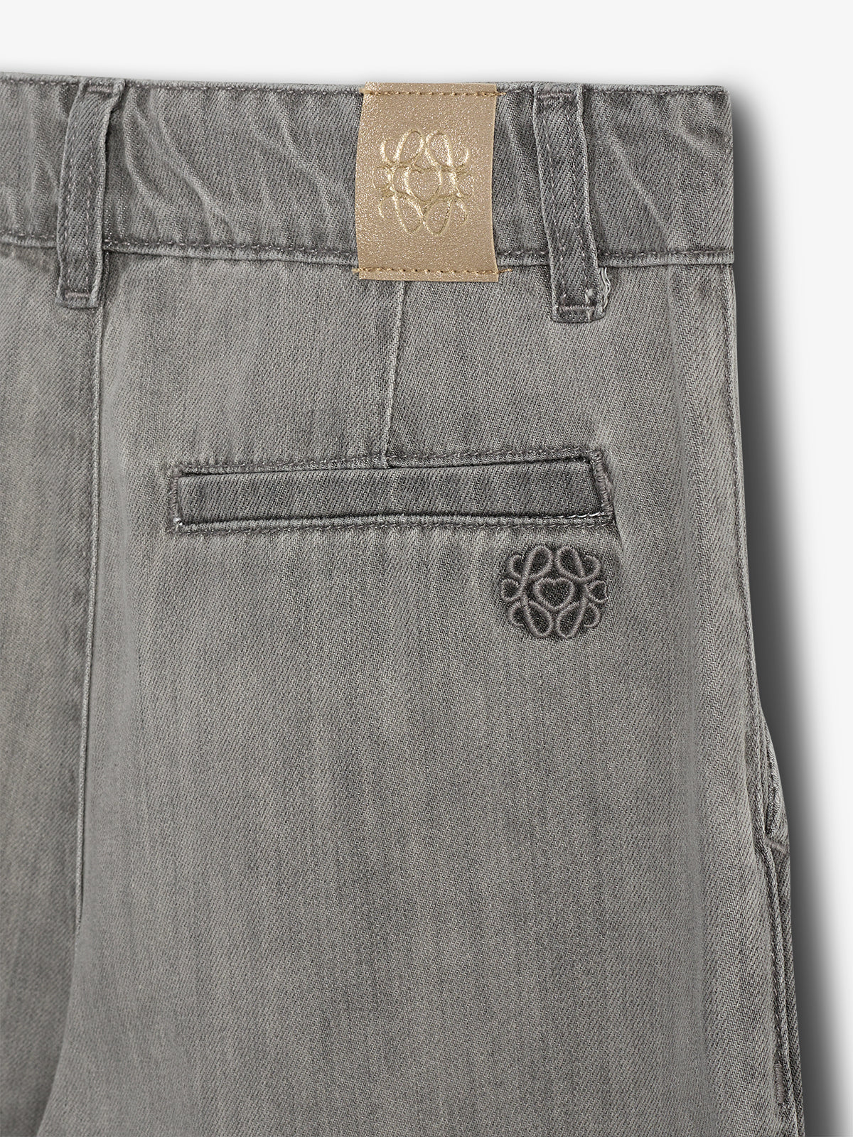 Gray denim pants display a beige leather patch and embroidered logo above a back pocket. Shadows suggest a studio setting.