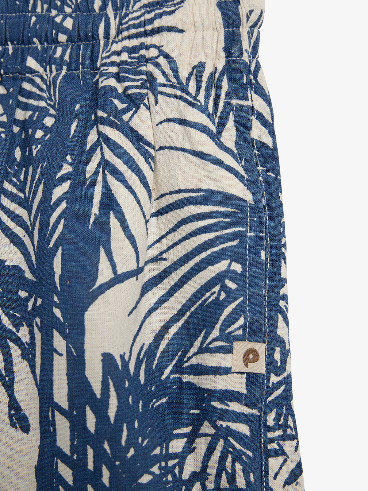 Blue and beige fabric with palm leaf pattern. The fabric forms part of a garment with an elastic waistband. A small tag with a logo is attached to the seam.