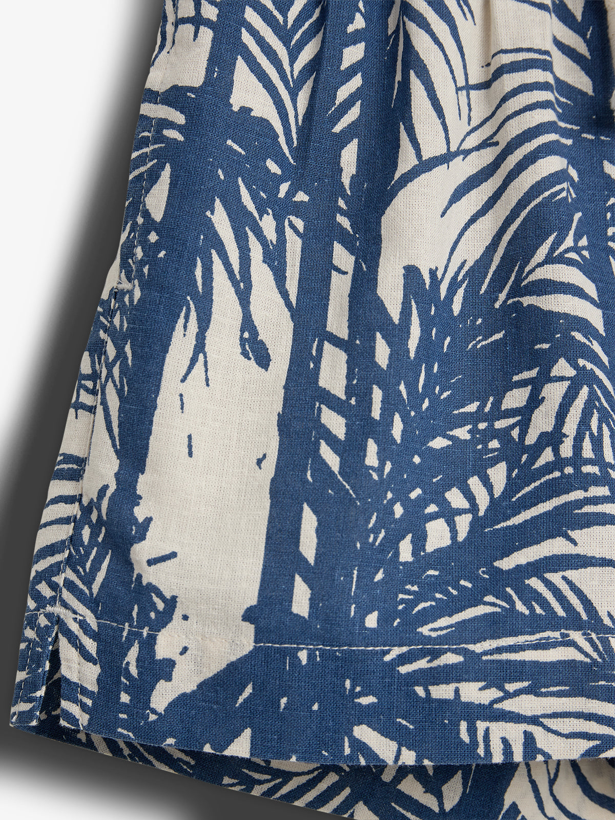 Blue palm tree silhouettes printed on white fabric, creating a tropical pattern. The texture is woven, with shadows on the left enhancing depth and dimension. No text is present.