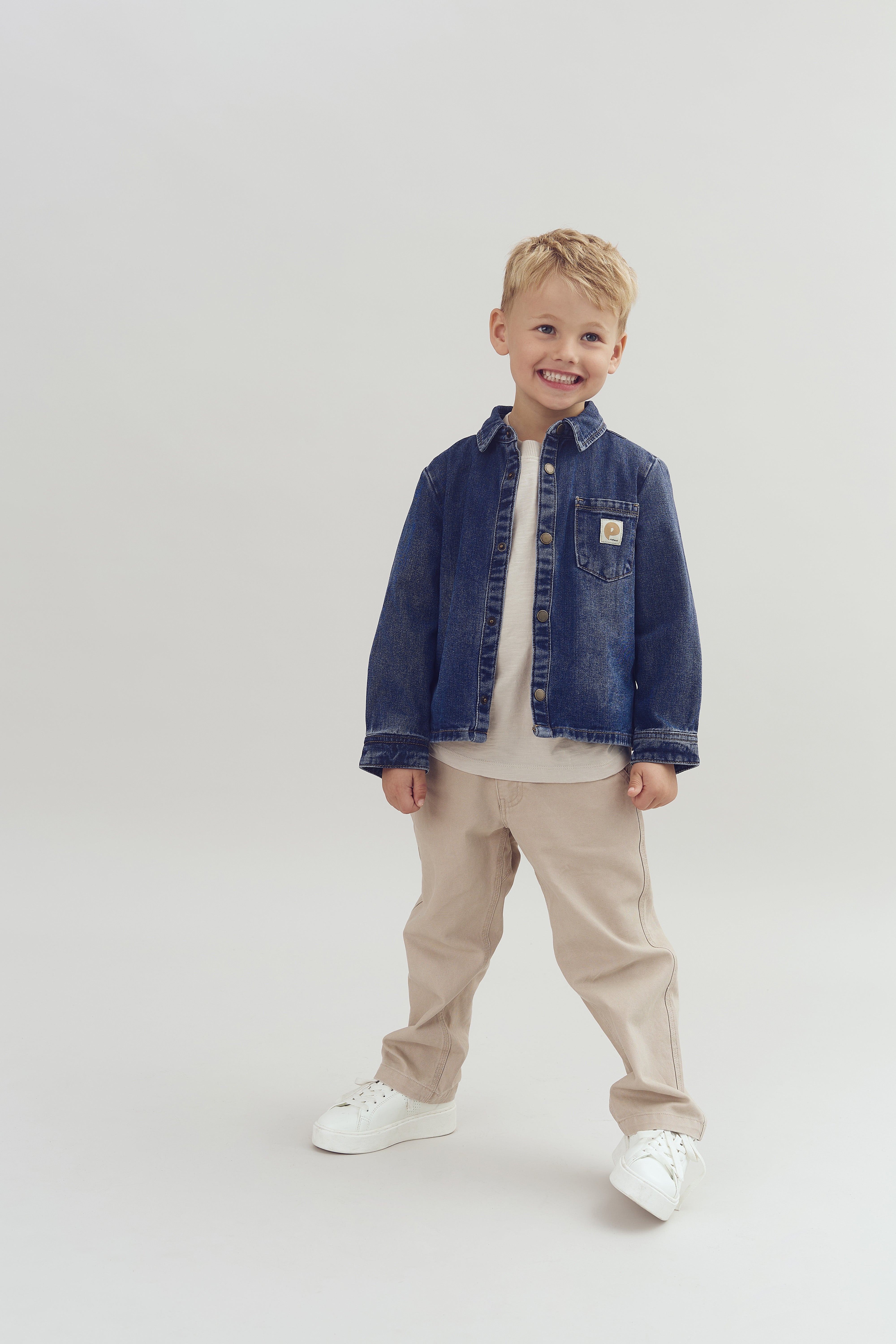 A smiling child stands confidently, wearing a denim jacket and beige pants, on a plain, light-colored background.