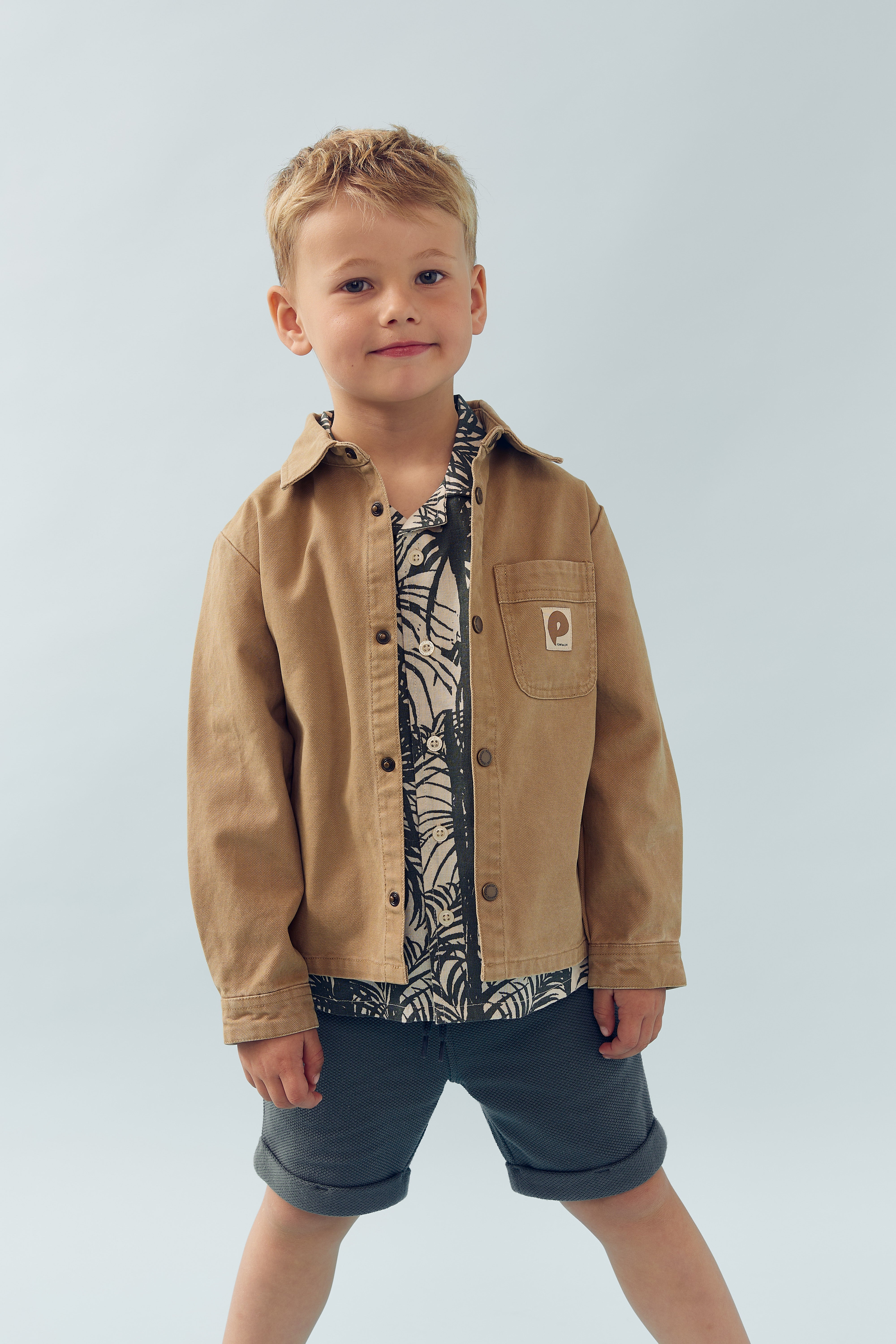 A young child stands confidently, wearing a brown jacket over a patterned shirt and dark shorts, against a plain light background.