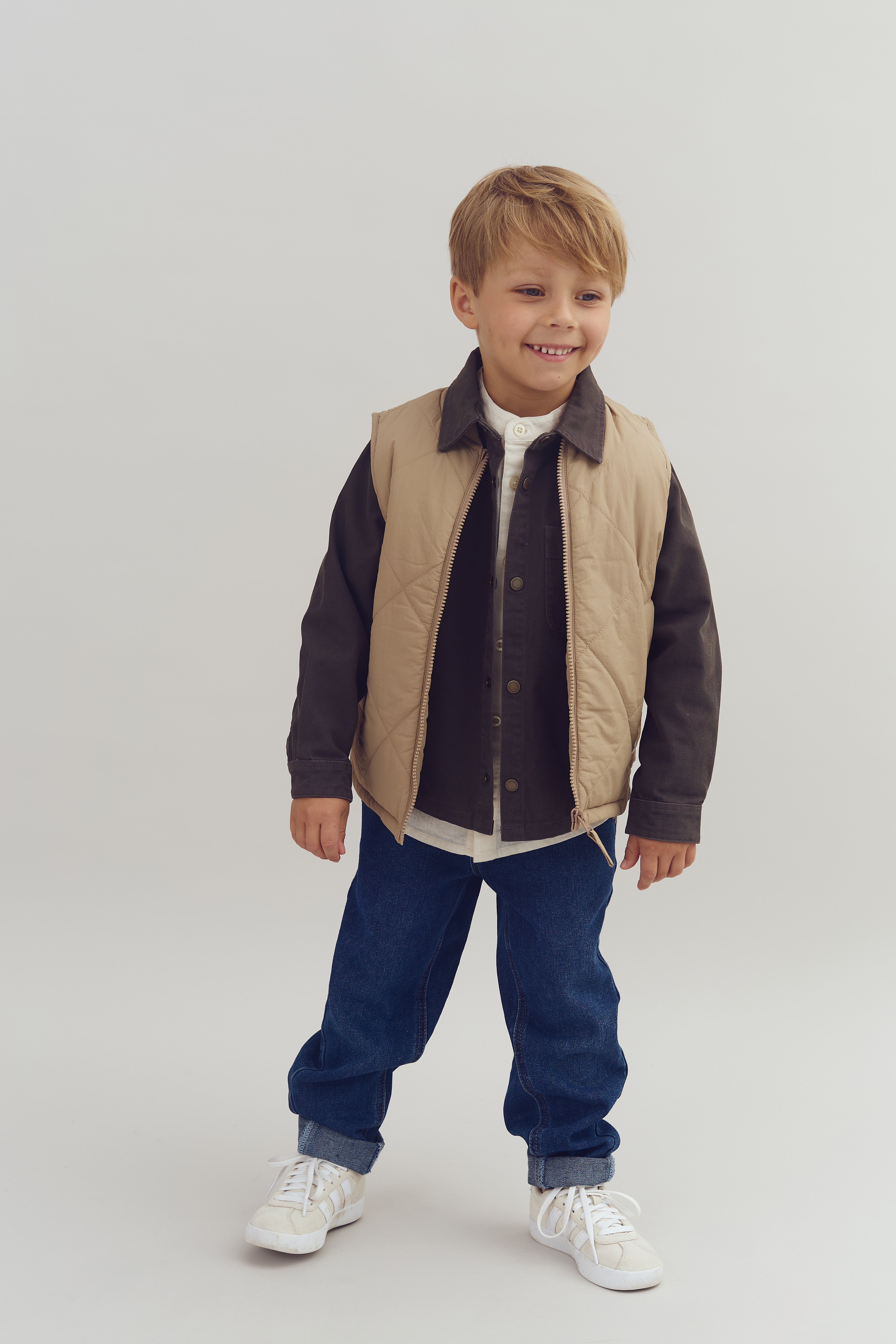 A smiling child stands confidently wearing a beige vest, dark shirt, blue jeans, and white sneakers against a plain light background.