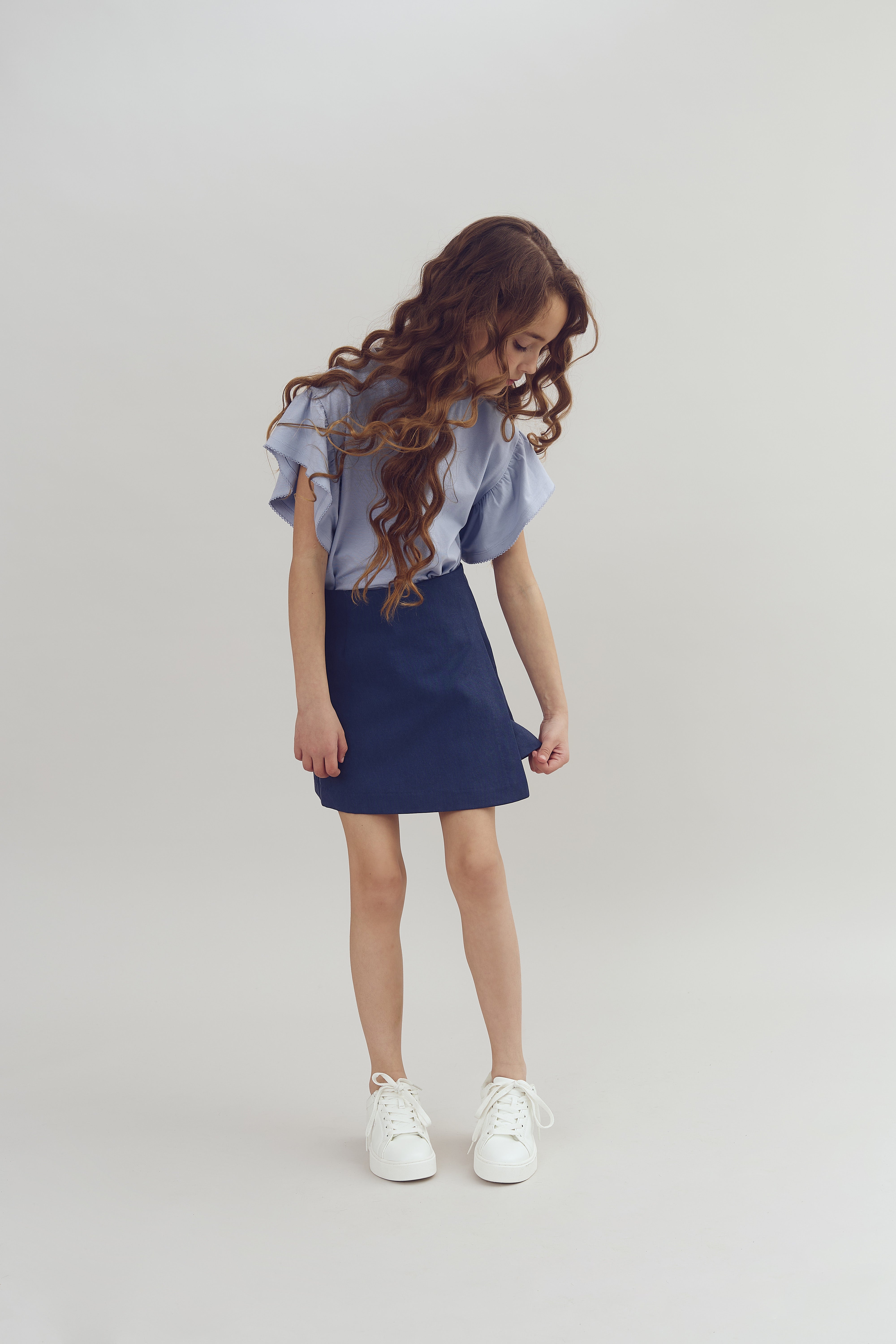 Girl in a blue top and skirt looks down, adjusting her skirt. She stands in a neutral, light-colored studio setting.