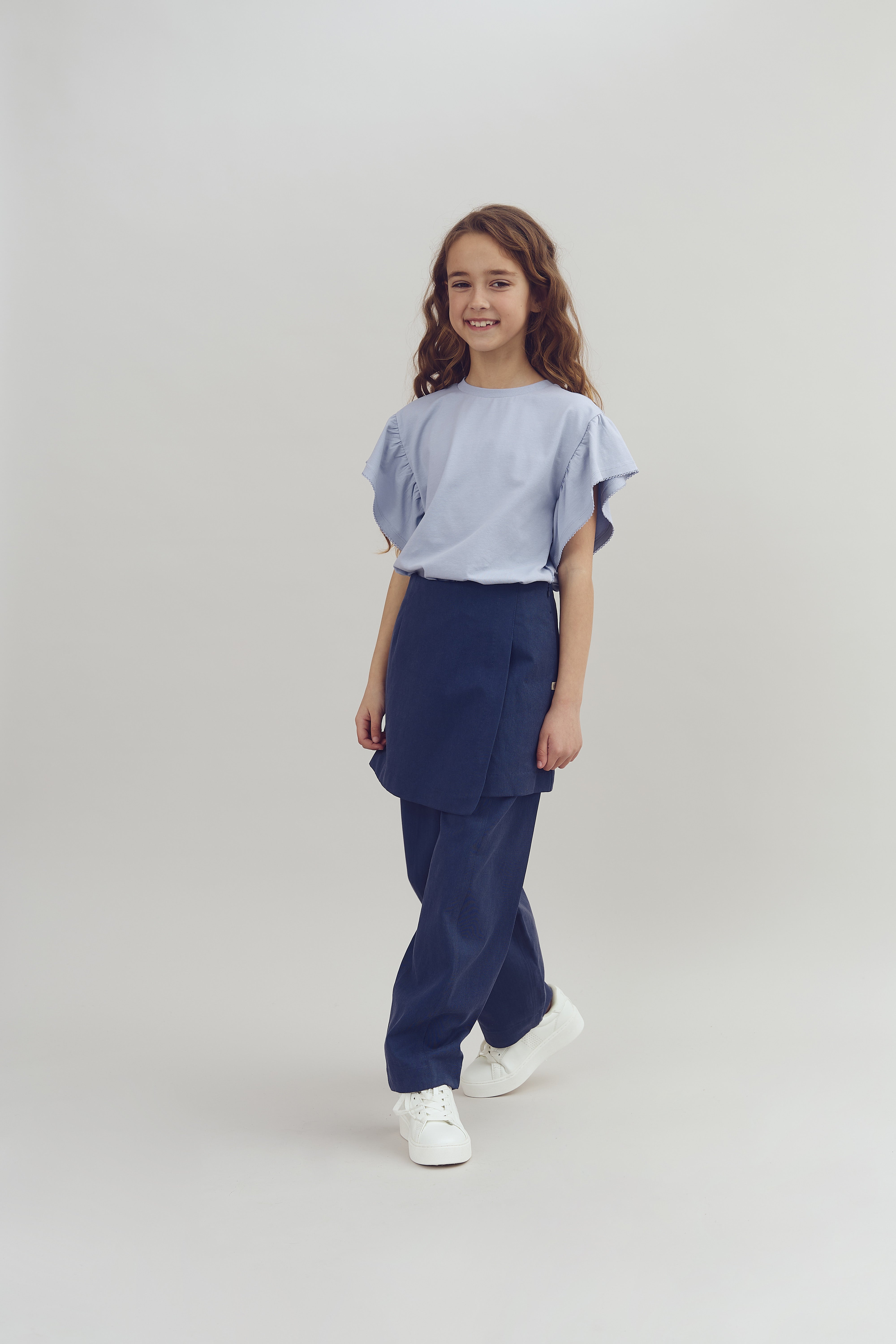 A young girl stands smiling, wearing a light blue top and dark blue pants with white shoes, in a plain, well-lit studio setting.