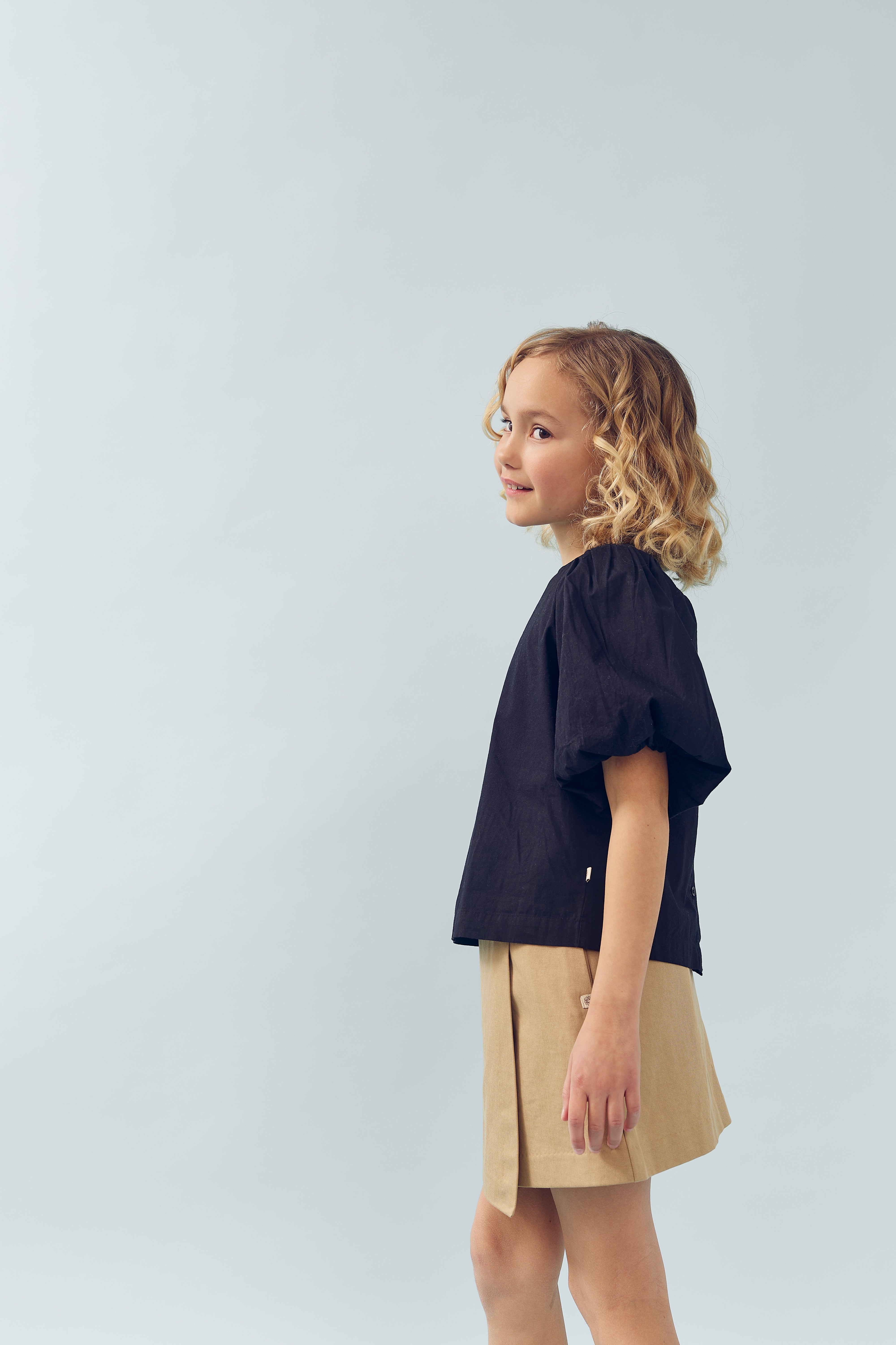 A young child with curly blonde hair stands smiling in profile, wearing a black top and beige skirt, against a plain light background.