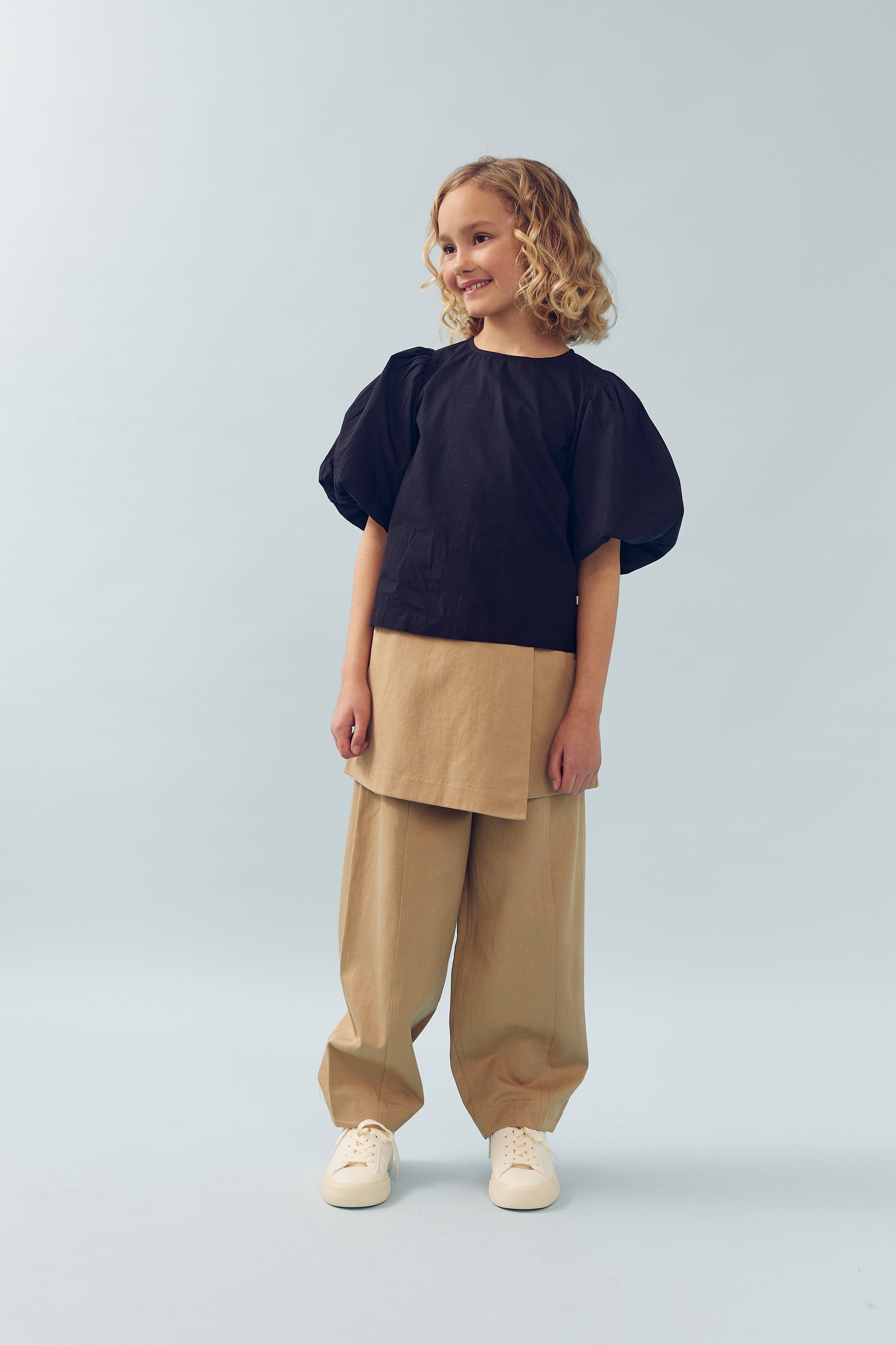 Child standing, wearing a black puff-sleeve top and tan pants, smiling and looking sideways. The background is a plain, light grayish-blue.