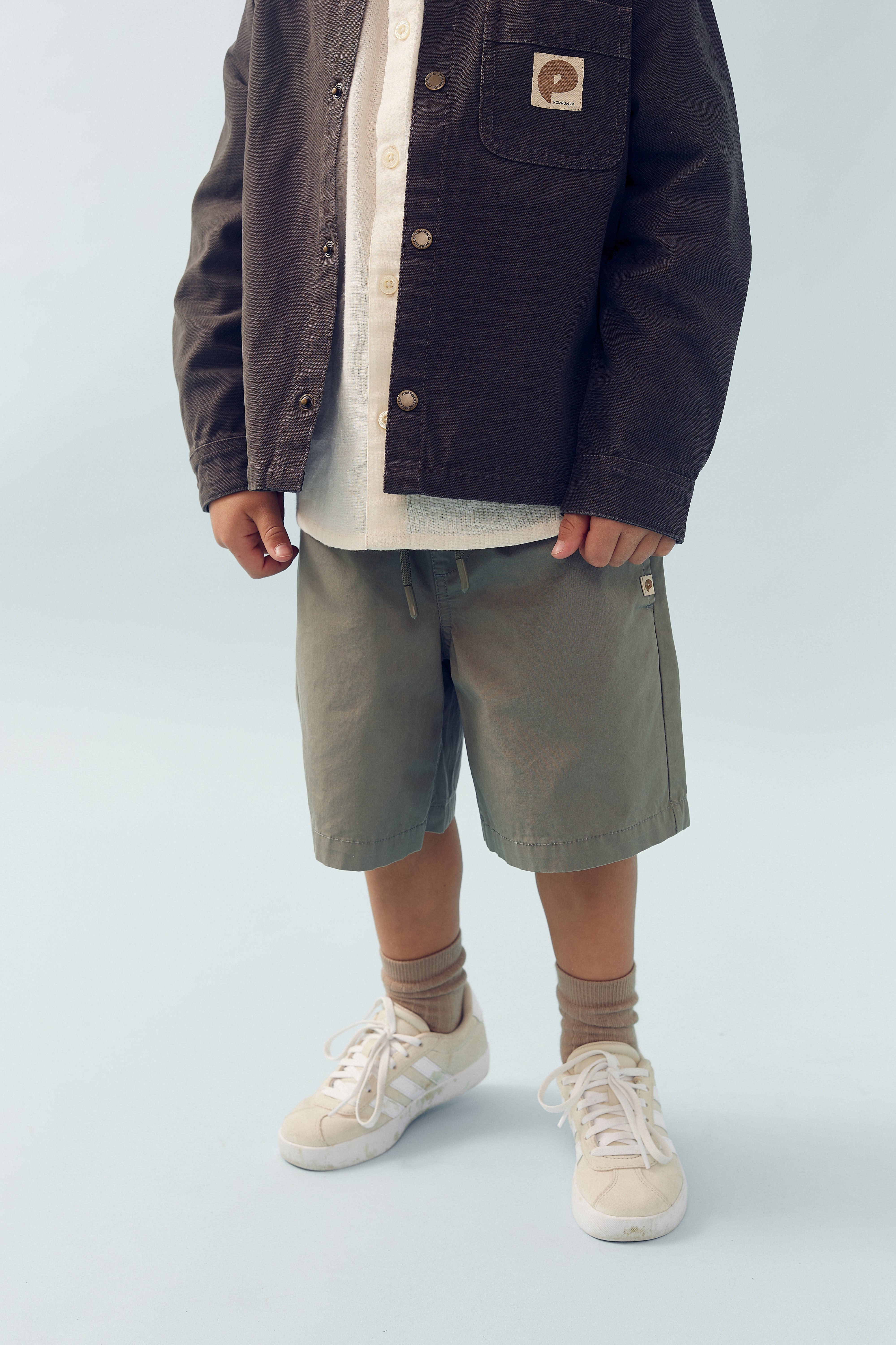 Child wearing a dark jacket with a logo, buttoned shirt, khaki shorts, and beige sneakers, standing with feet apart against a light gray background. Logo reads "Pikul Ark."