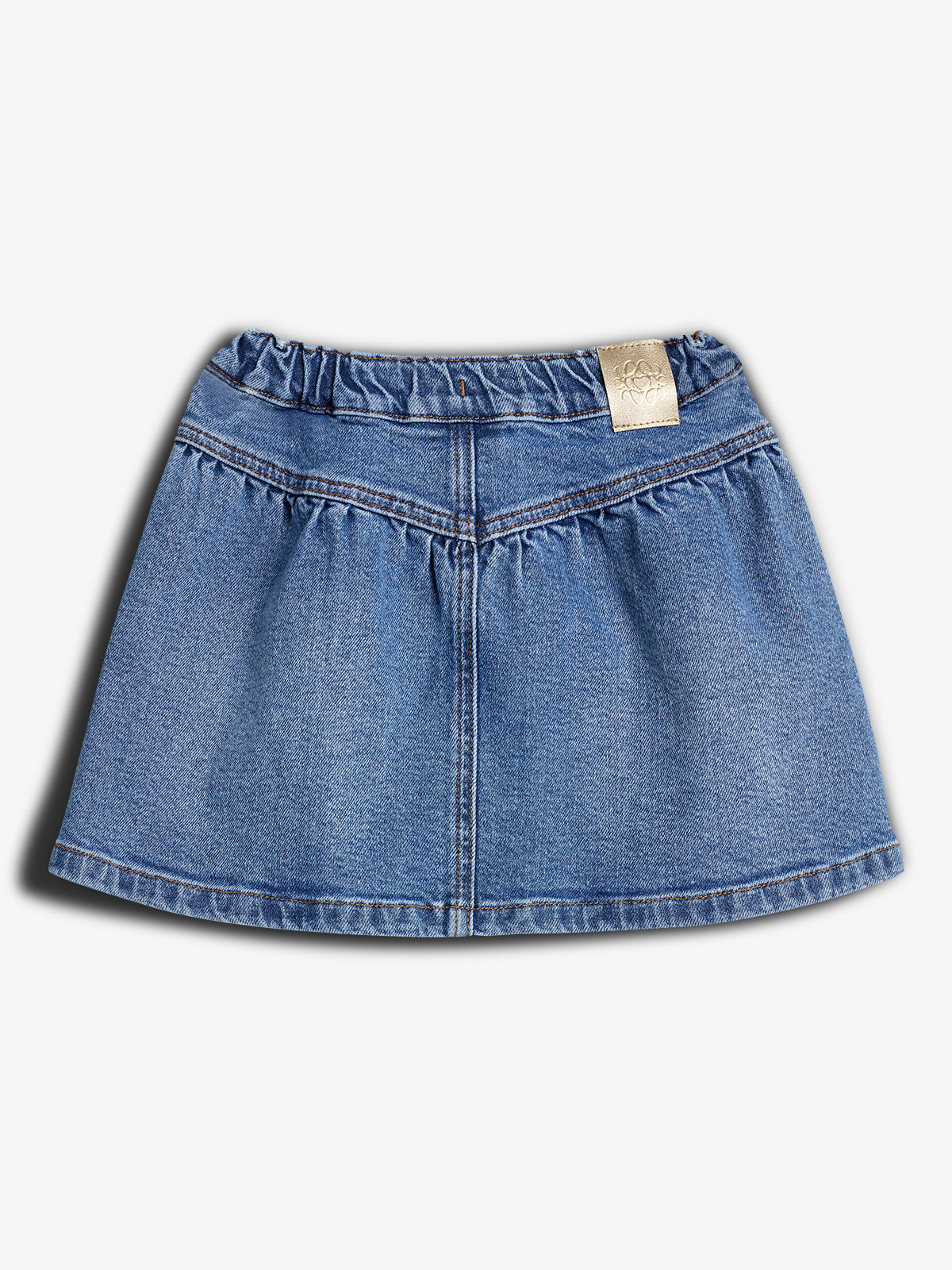 Denim skirt lying flat, displaying gathered waistband with a small beige tag featuring a logo. The background is plain white, emphasizing the skirt’s blue color and stitching.
