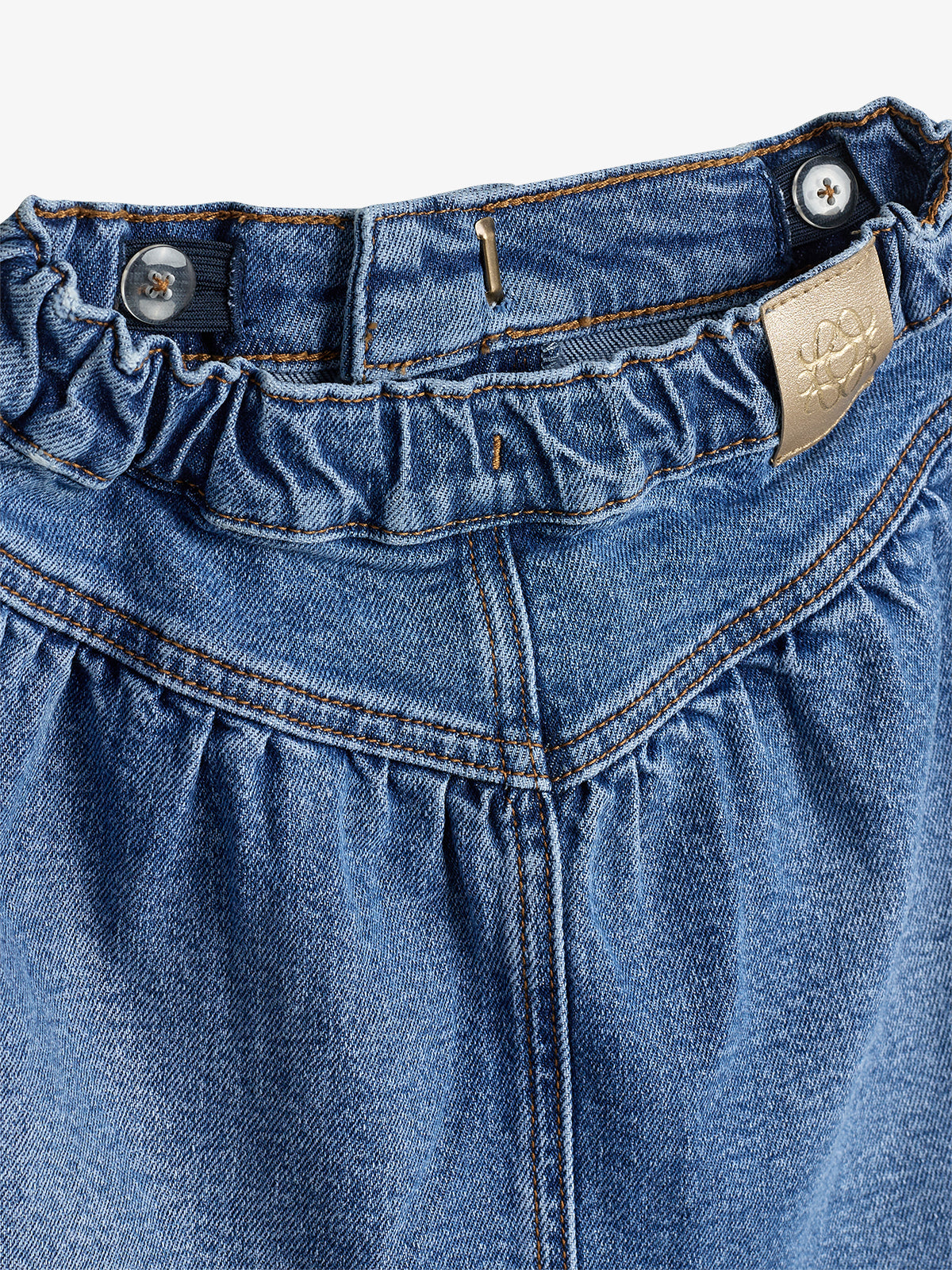 Denim skirt with an elastic waistband and two buttons is displayed in close-up. A tan label featuring a floral design is sewn on.