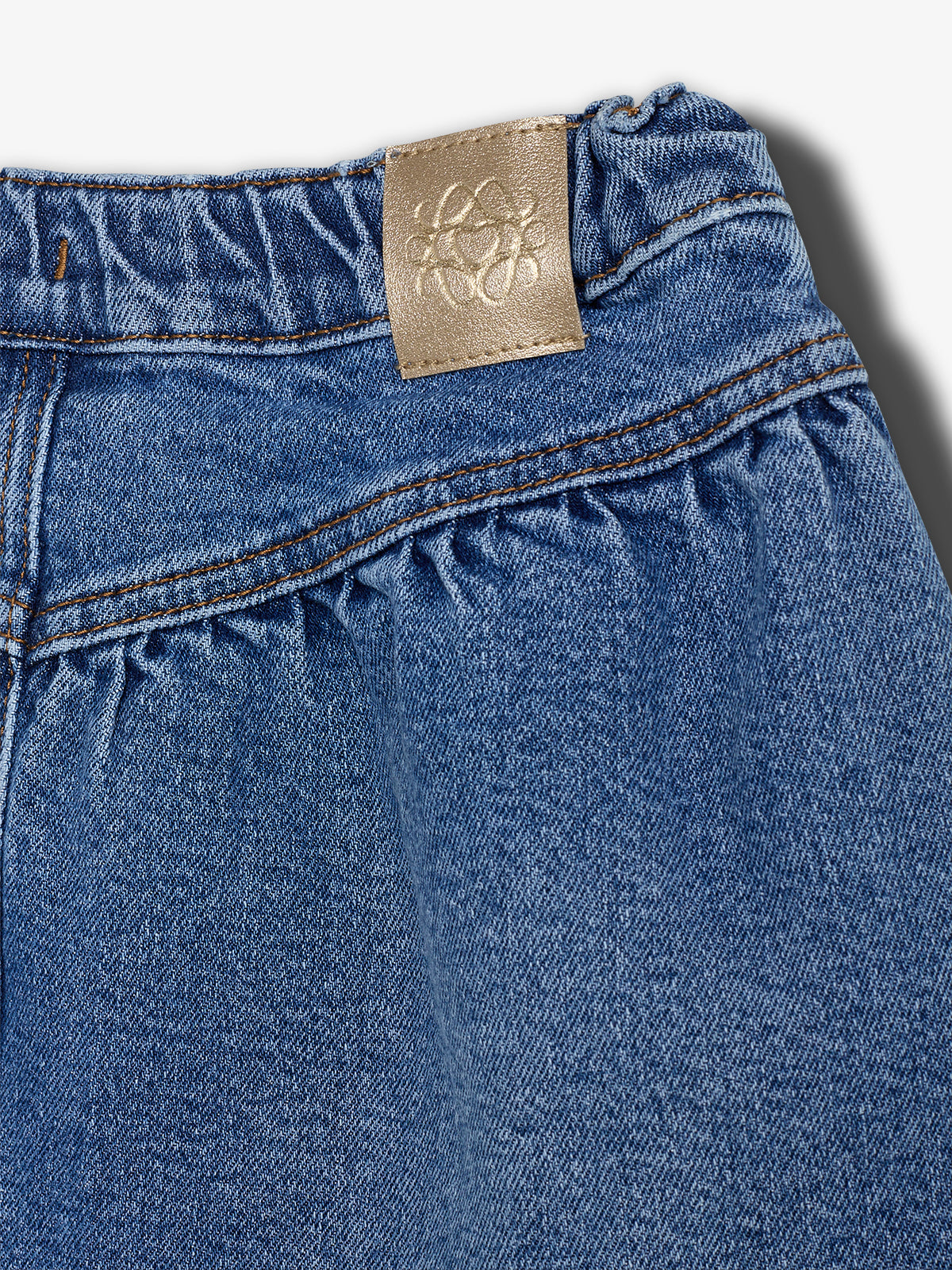Denim fabric features gathered stitching with a beige leather patch displaying an embossed design, set against a plain white background.