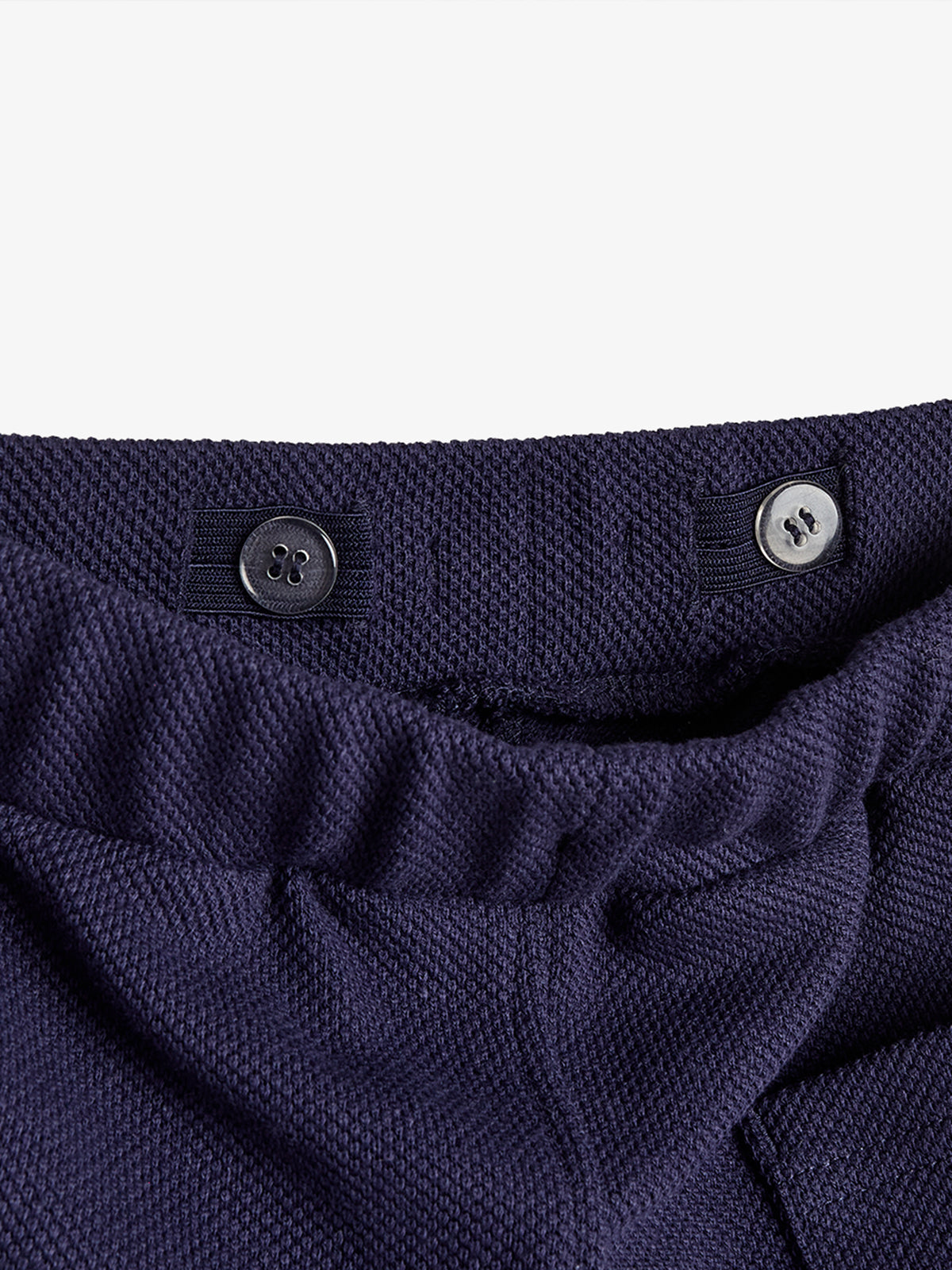 Navy blue textured pants feature an adjustable waistband with a black button for fastening. The background is plain white, highlighting the clothing detail and construction.
