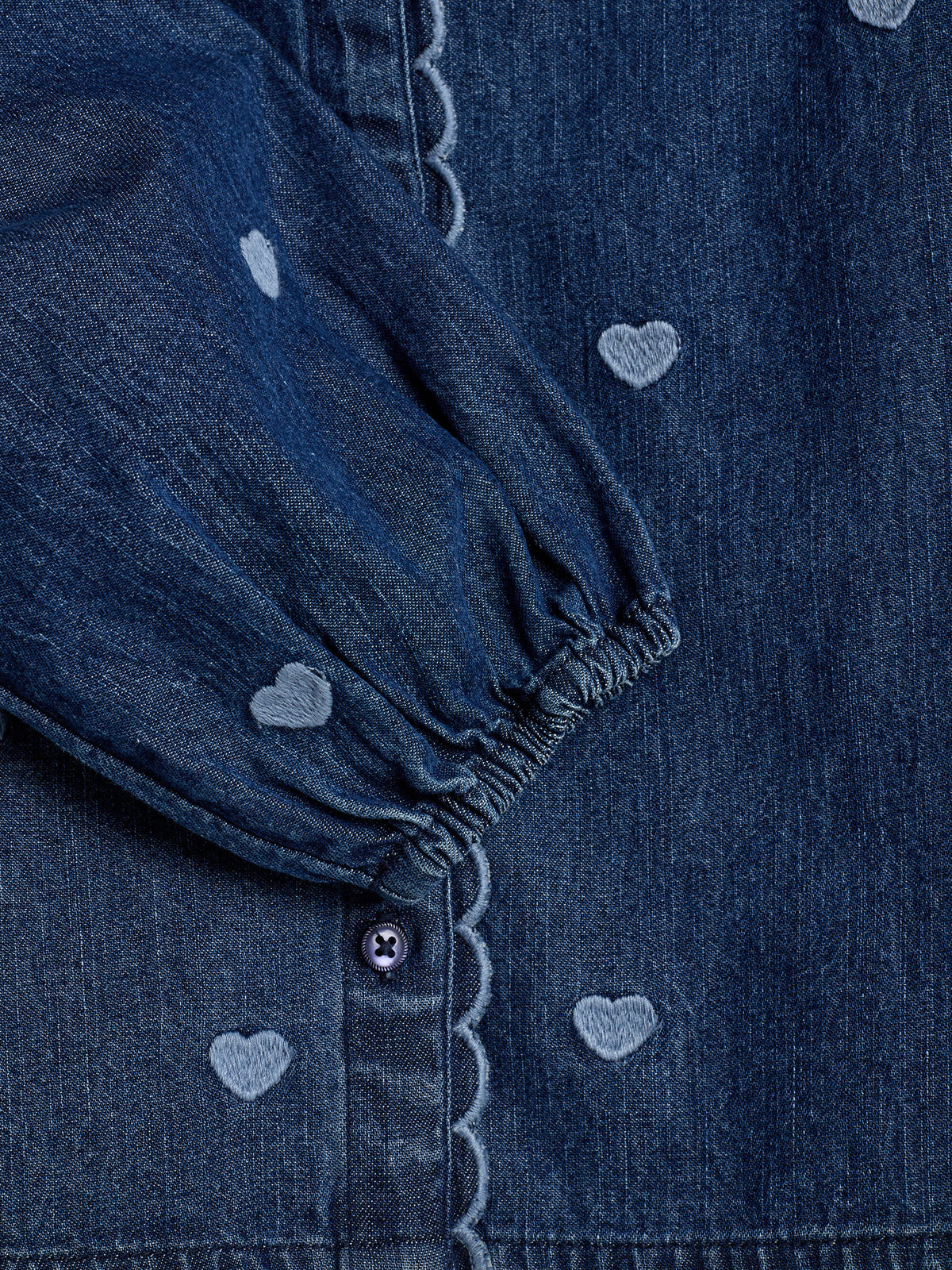 A dark denim fabric with small embroidered heart patterns, featuring a gathered sleeve close to a buttoned seam with scalloped edges, creating a textured design.