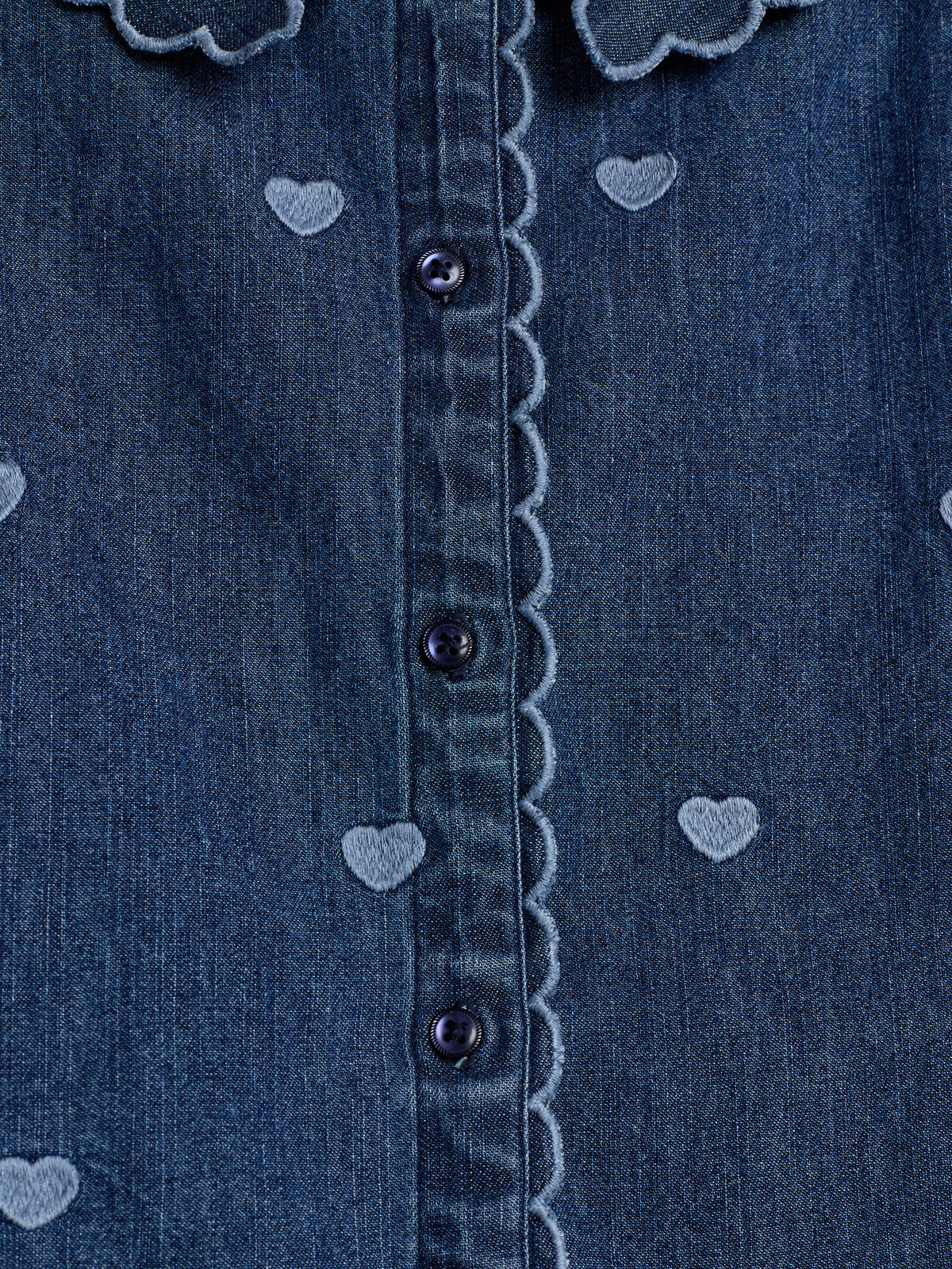 Denim fabric with small embroidered heart patterns decorates a shirt or dress featuring dark buttons and scalloped edges, suggesting a structured, stylish garment.