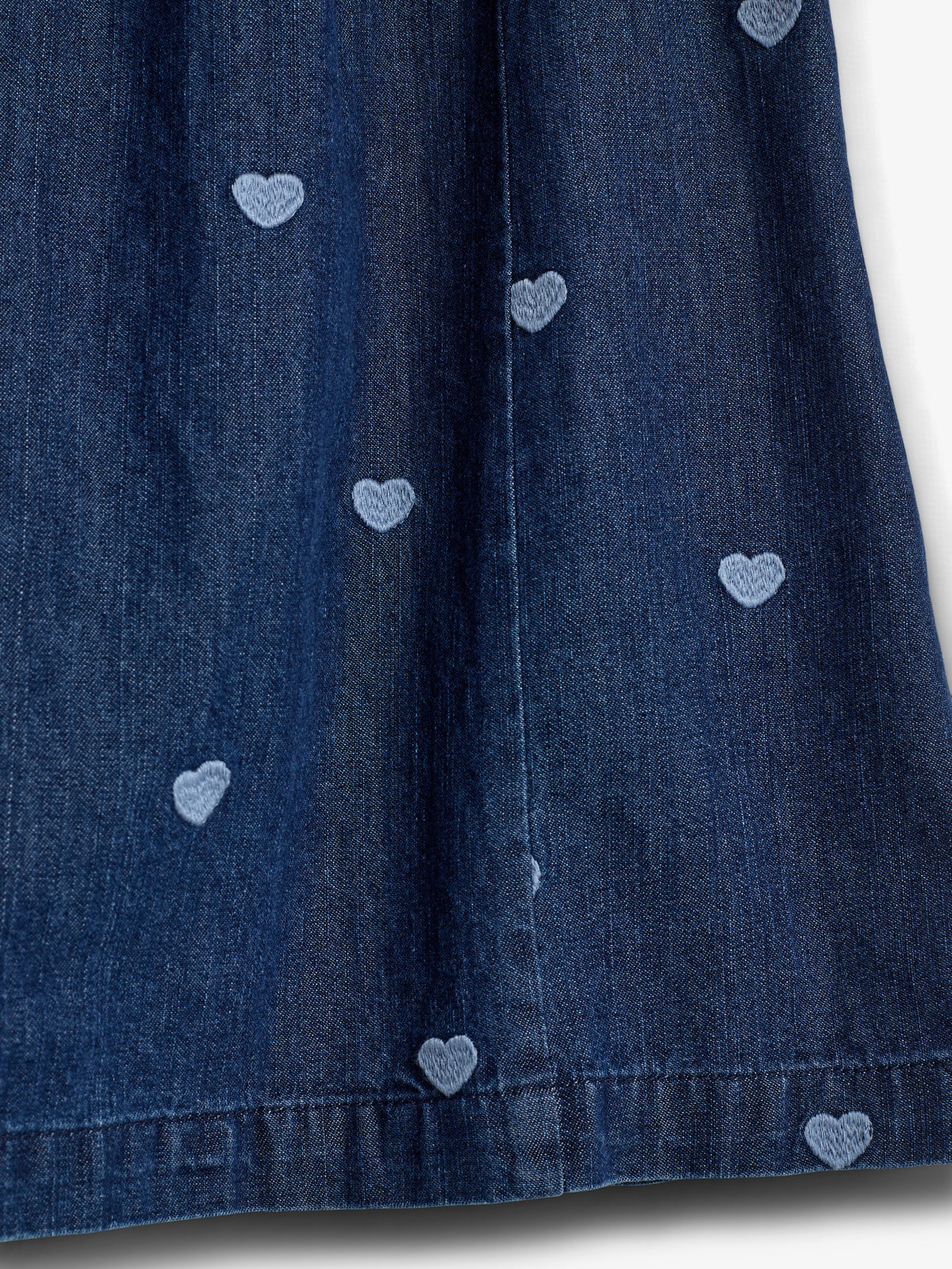Dark blue denim fabric is adorned with small, light blue embroidered heart patterns, creating a playful design. The fabric appears to be part of a garment, set against a plain background.