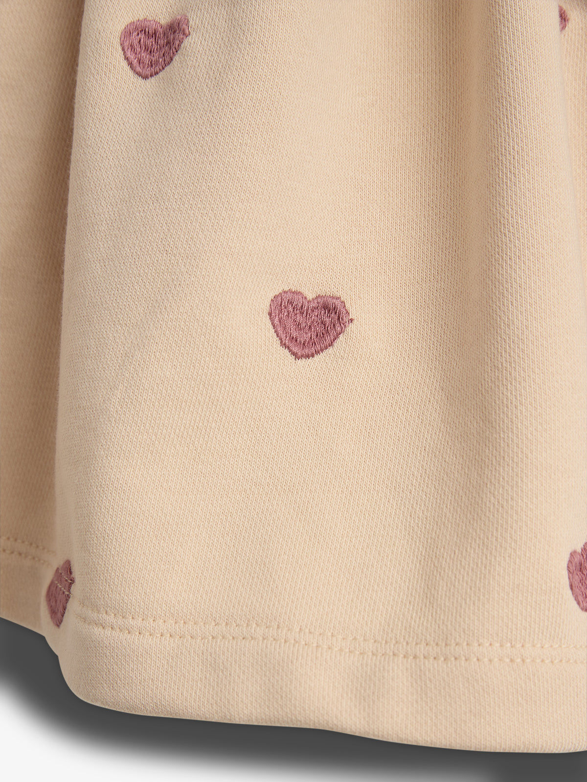 Beige fabric features small, embroidered pink hearts, creating a decorative pattern. The cloth appears gathered and softly draped, suggesting a garment's hem.