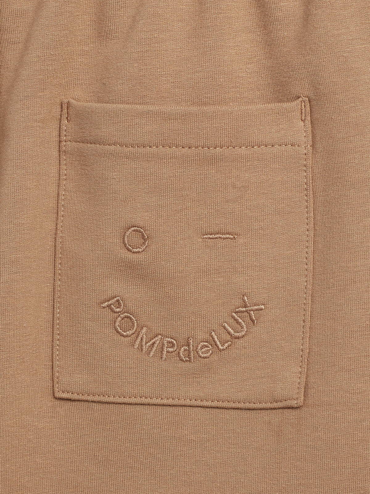 A beige fabric pocket features embroidered text and a simplistic face design. The text reads "POMPdeLUX." The pocket is sewn onto a matching fabric, blending seamlessly with the background.