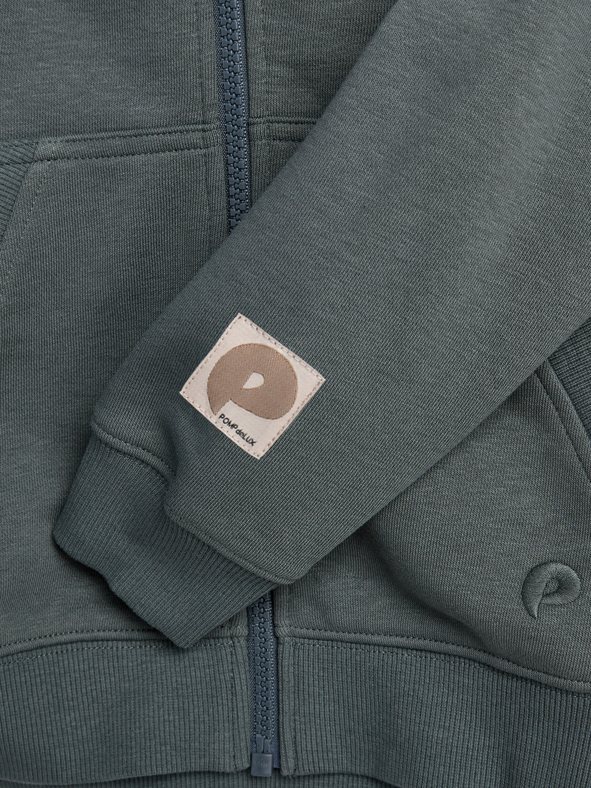 A green zippered jacket with a sleeve featuring a beige patch displaying a brown circle and the text "polar skate" underneath. The jacket is folded neatly.