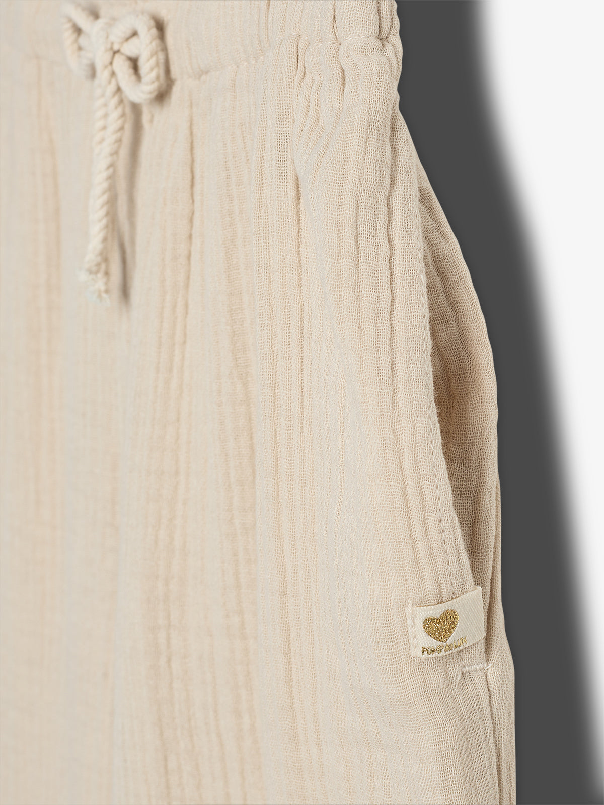 Beige pants hang with a drawstring at the waist; fabric is textured with vertical lines. A label near the pocket features a gold heart with text: "POUPETTE PARIS."