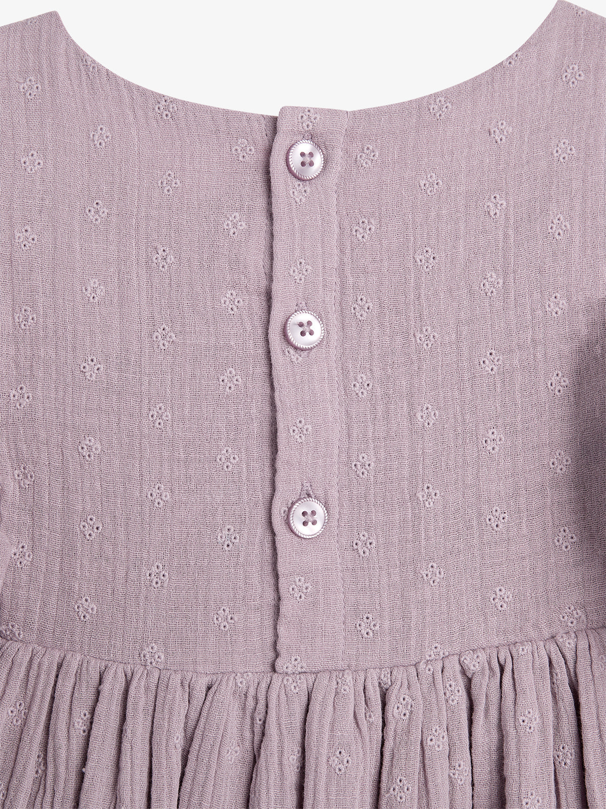 A textured, lilac fabric dress with small embroidered patterns features three vertically aligned buttons. The dress has a gathered waist, creating soft pleats below the buttoned section.