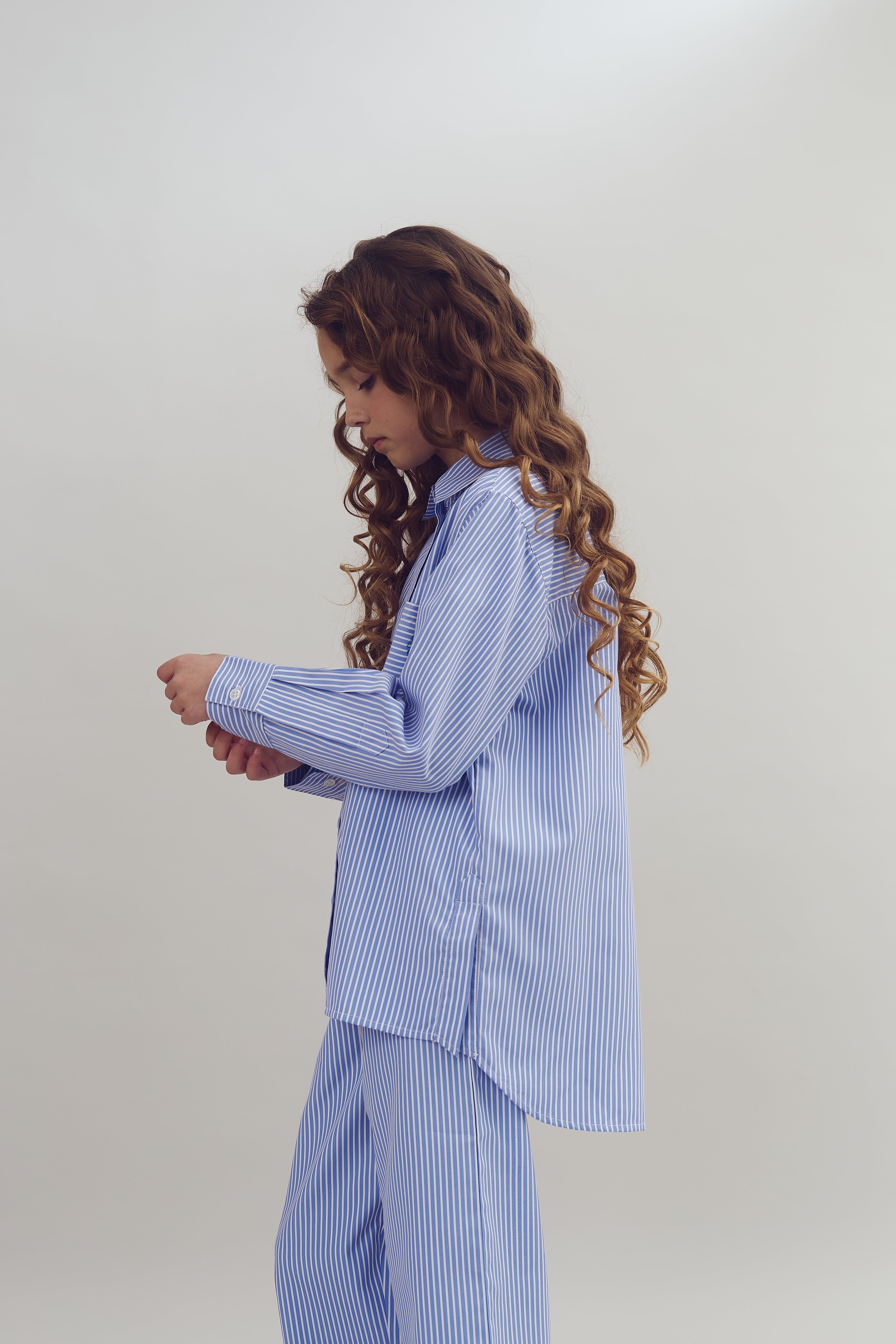 A person in blue striped pajamas looks down while adjusting the sleeve. The background is plain and neutral, giving a minimalistic feel.