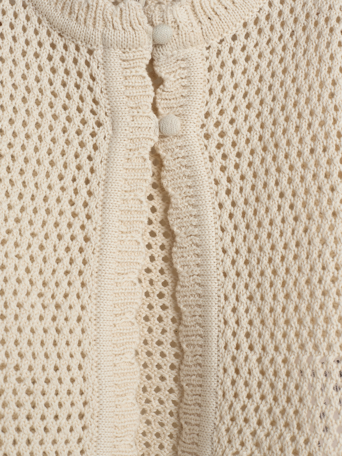 A beige knitted fabric with a textured, open-weave pattern features a button-down closure with two fabric-covered buttons in the center, surrounded by a ribbed edge.