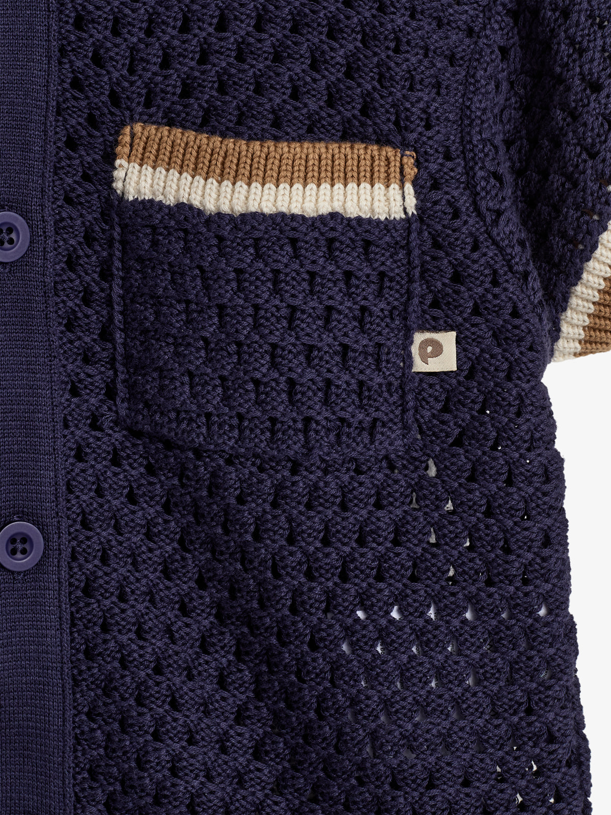 A dark purple crochet cardigan features a brown and white striped pocket. A small label with a stylized “p” is near the pocket, surrounded by a textured fabric pattern.