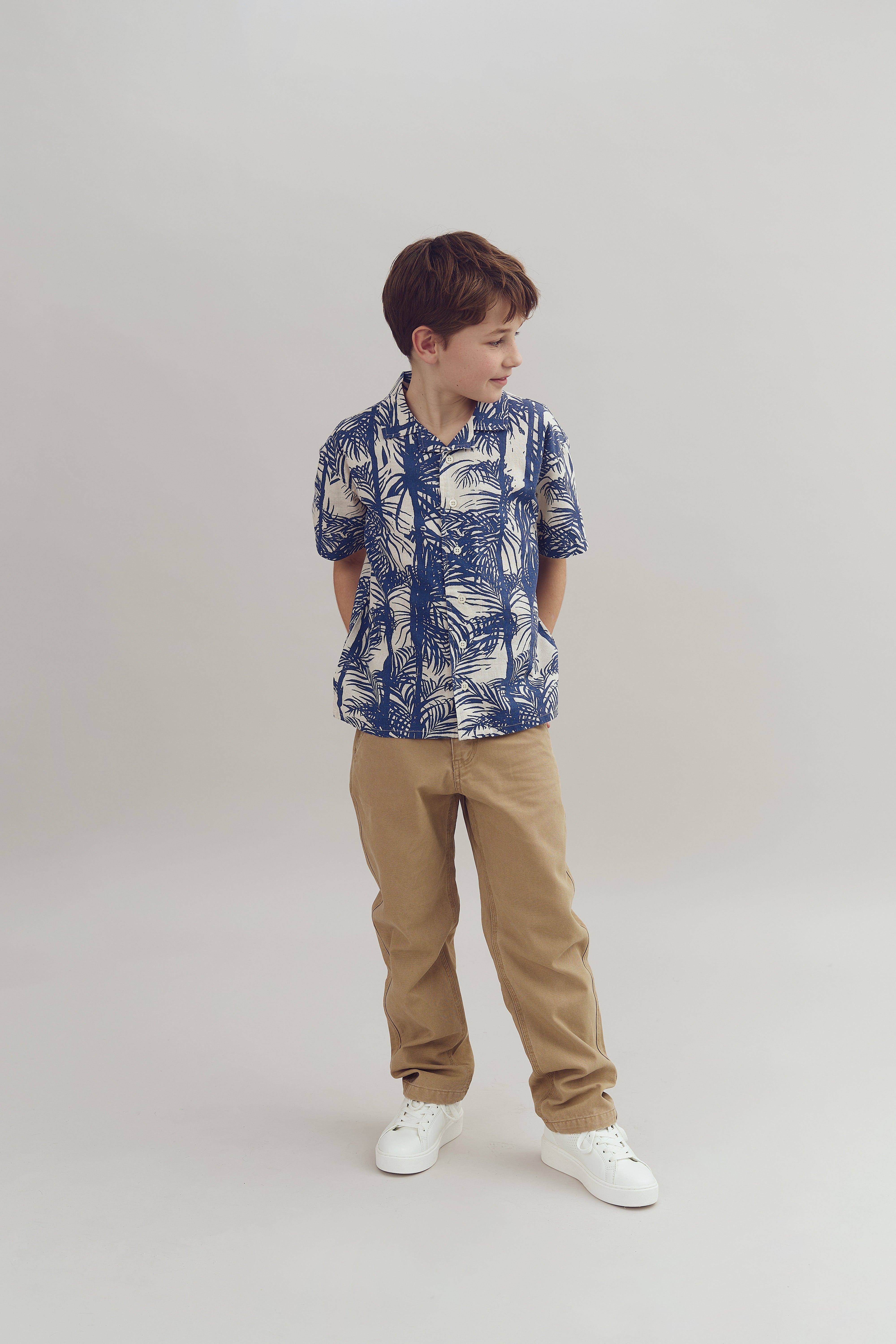 Ein Junge steht, blickt zur Seite; er trägt ein blau-weiß gemustertes Hemd und beige Hosen. Der Hintergrund ist neutral und schlicht.