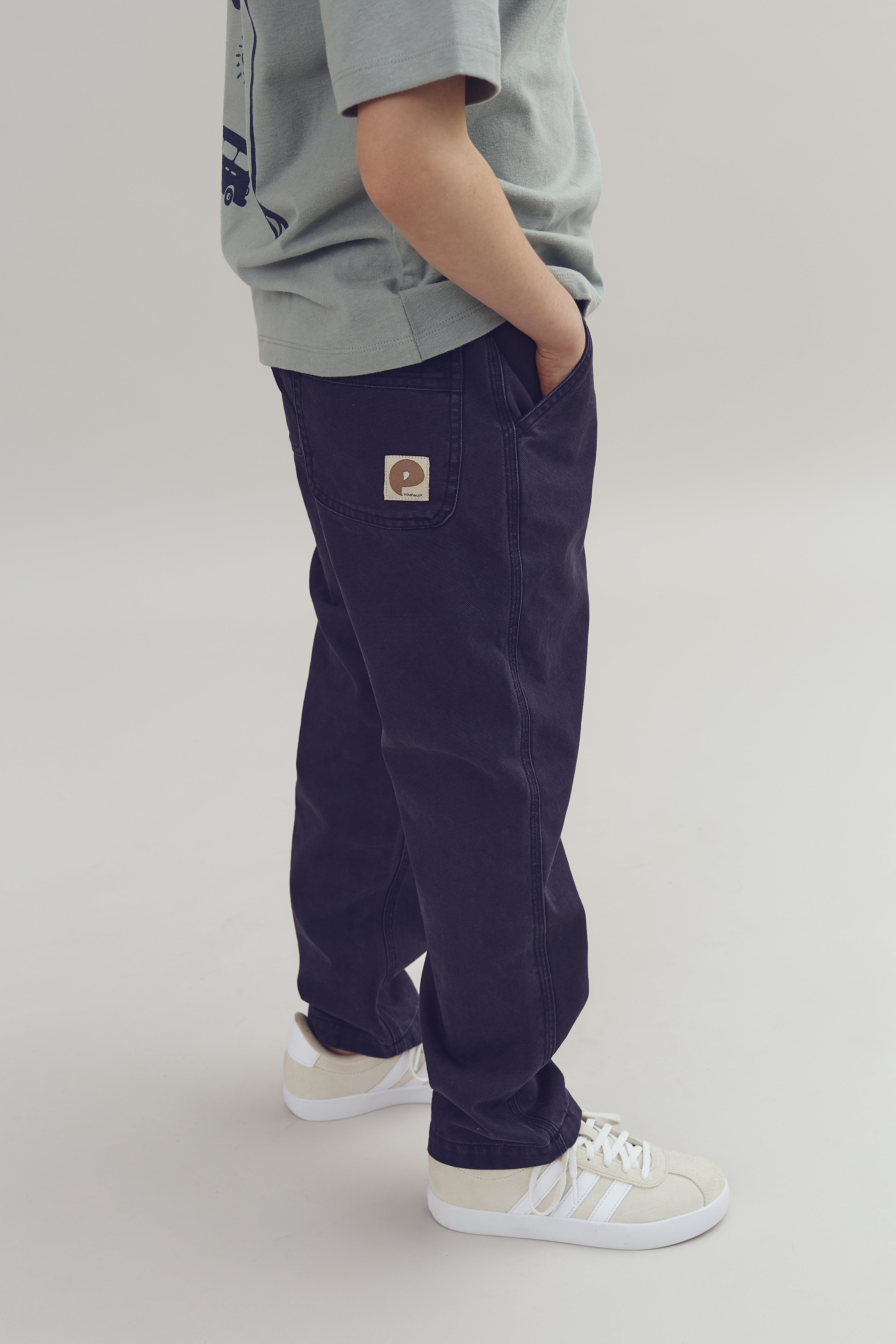Eine Person trägt blaue Jeans und weiße Turnschuhe; die Hände sind in die Hosentaschen gesteckt. Auf der Jeans ist ein Etikett mit einem Logo und dem Text „the animals“ zu sehen.