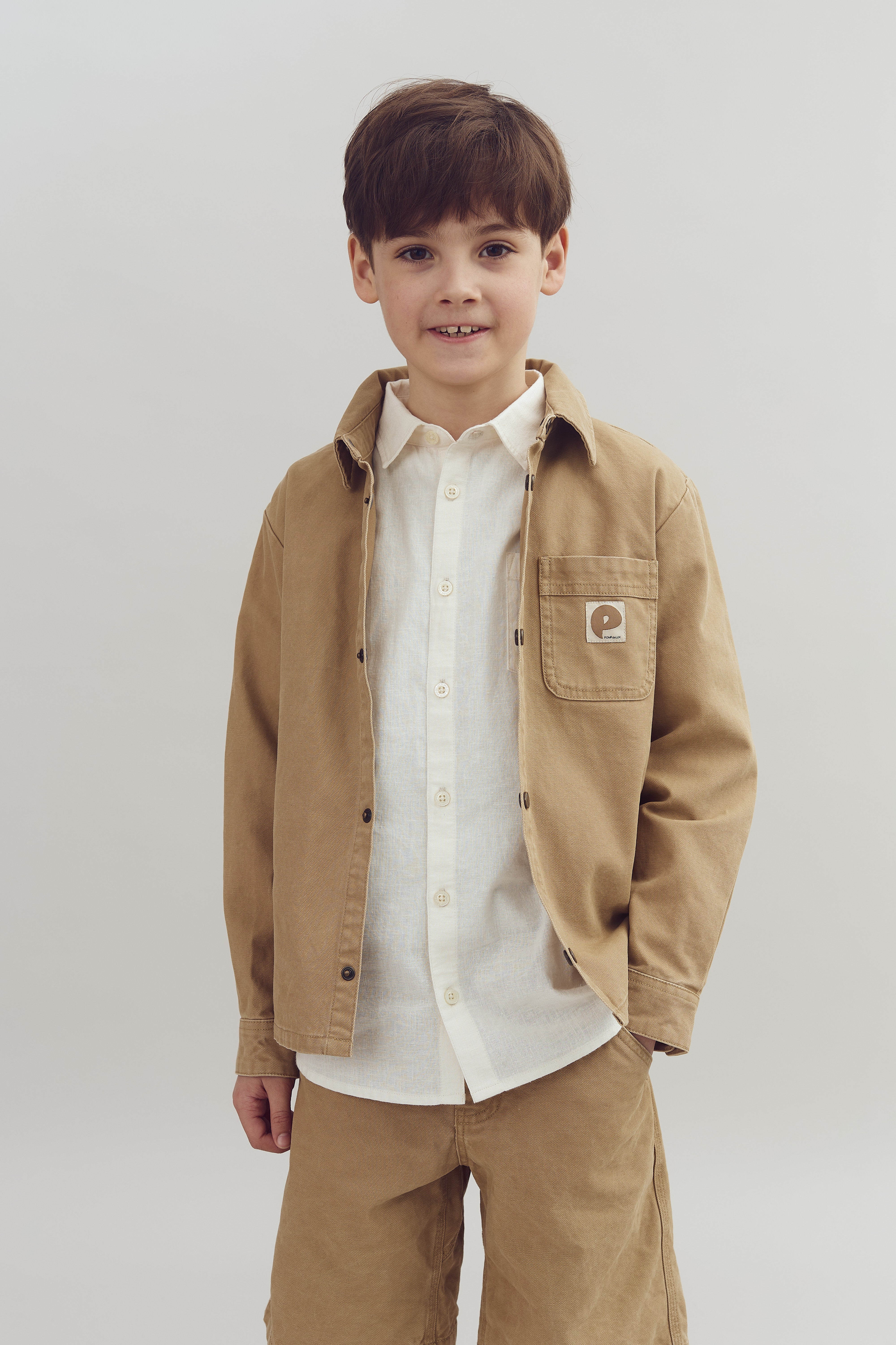 A child stands smiling, wearing a beige jacket with an embroidered "e" on the pocket and a white shirt. The background is plain and neutral.