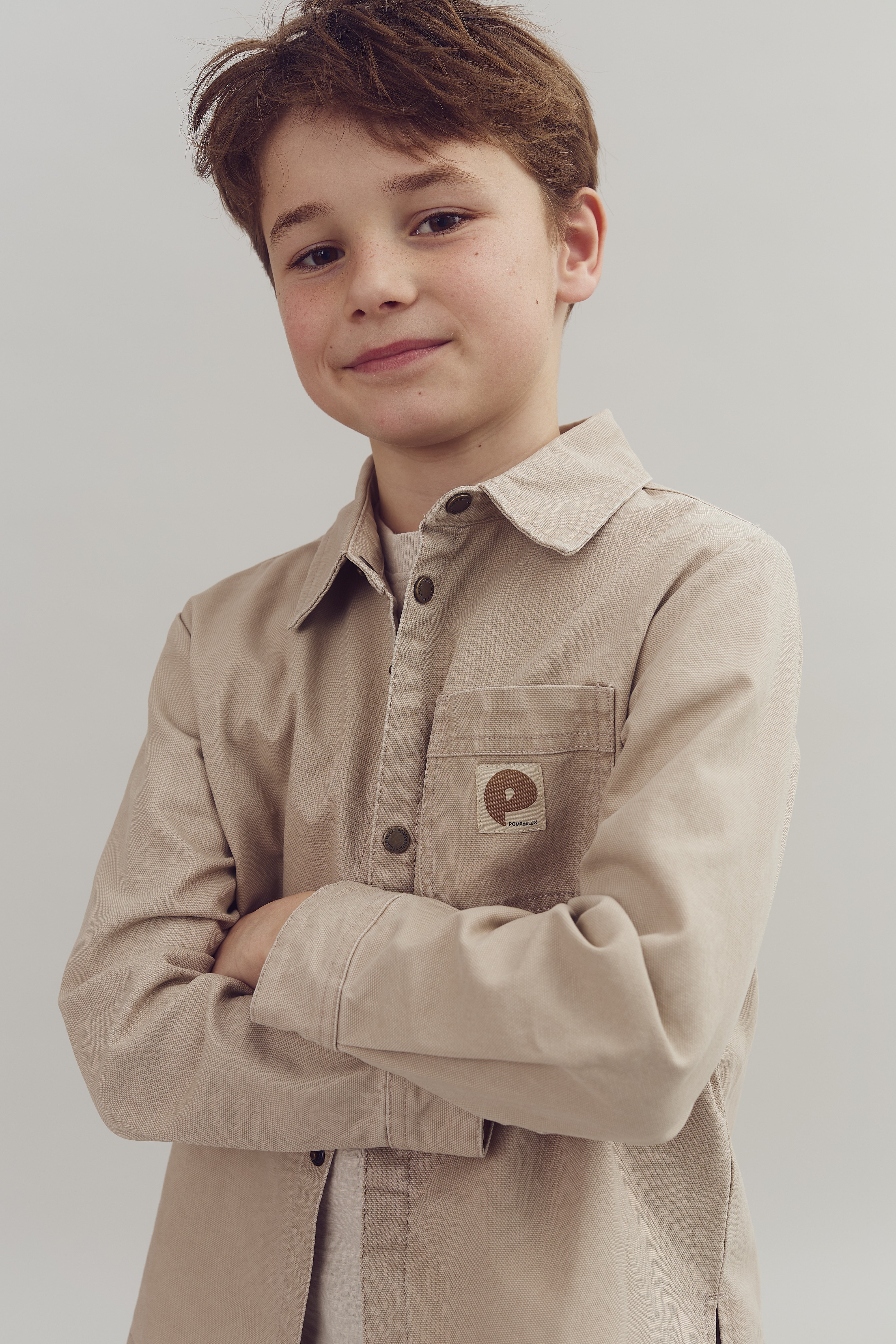 A child stands confidently, arms crossed in a beige jacket with a "P" logo on the pocket. The background is plain and neutral.